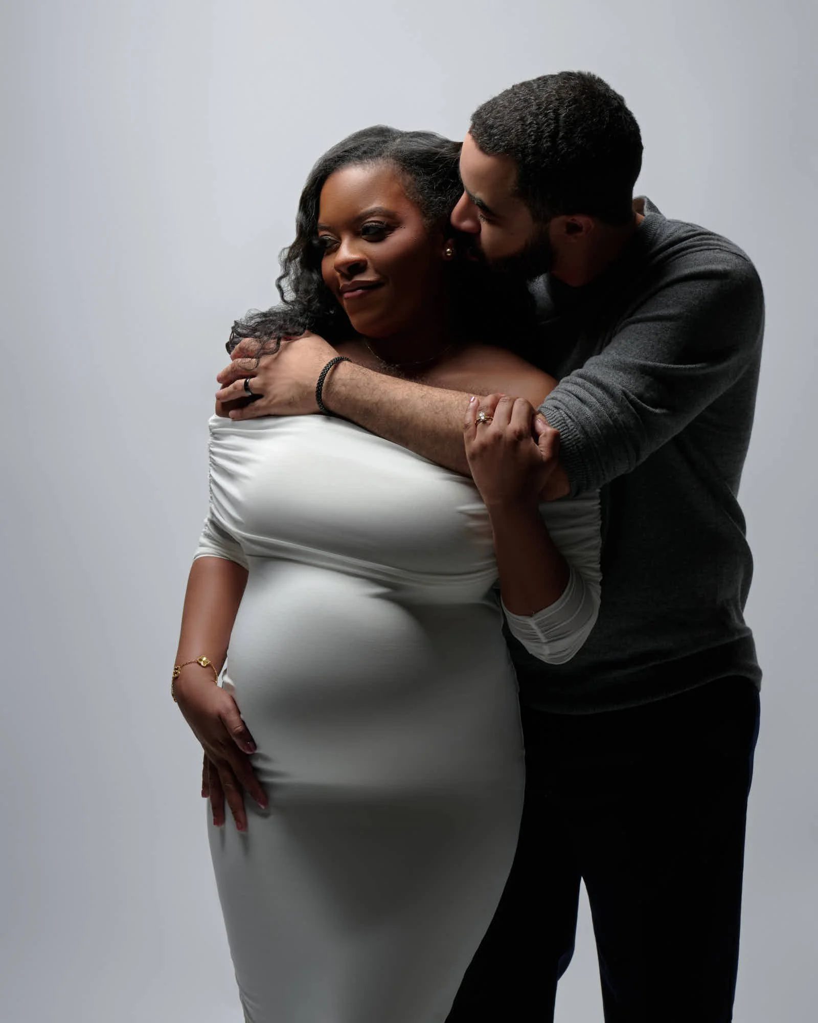 Partner wraps her in a hug from behind, her white dress hugging the bump.