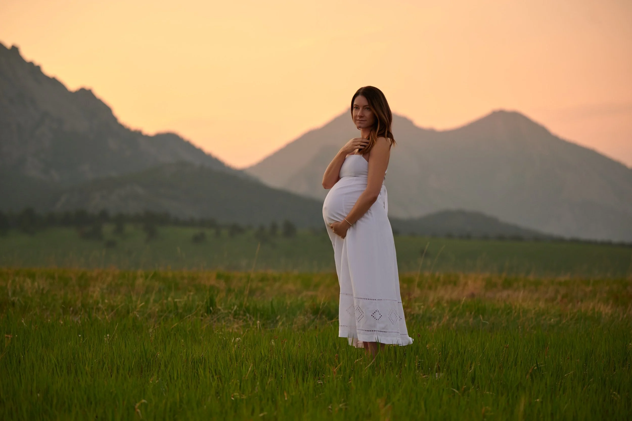 A pregnant woman standing in a field at sunset in a white dress in Colorado.