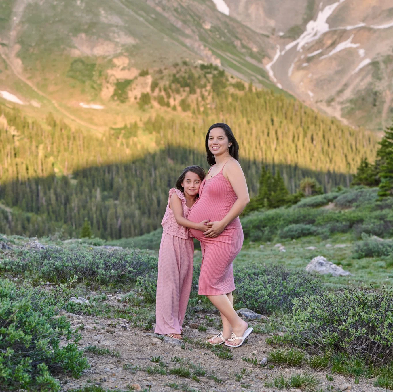 Mom and daughter hug in matching pink beneath a rugged alpine cirque.