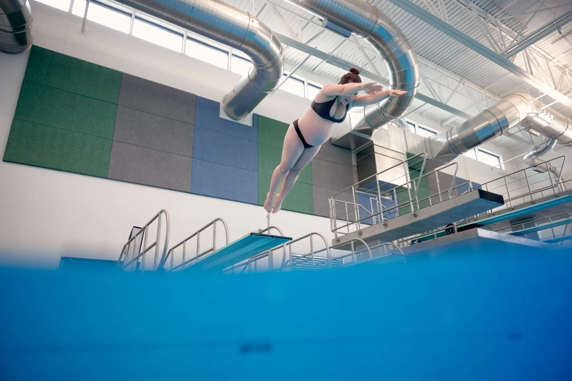 Springboard diver in flight above the pool, natatorium ramp and blue wall behind.