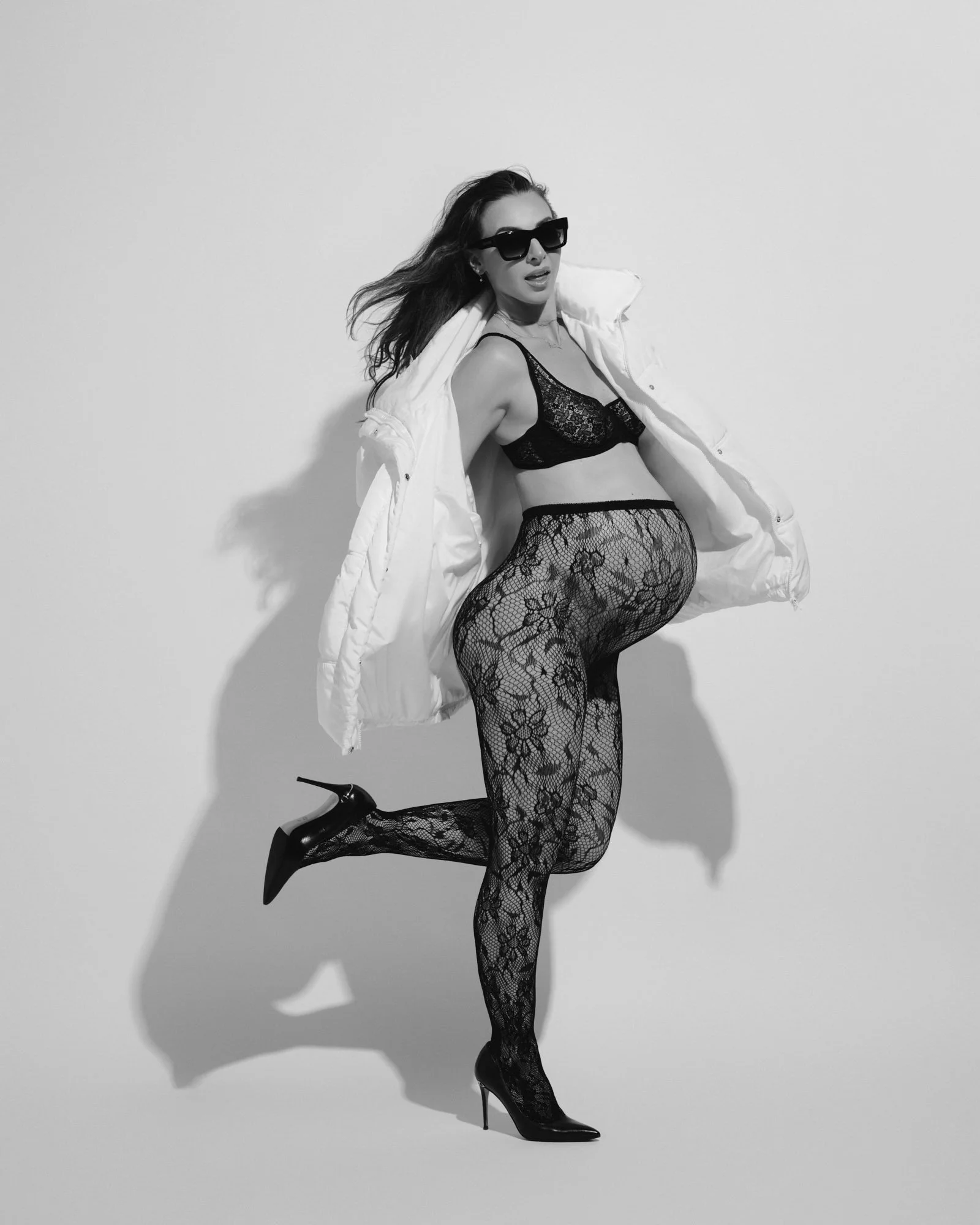 Black and white in a lace bodysuit and heels, one leg kicked back, sunglasses on.