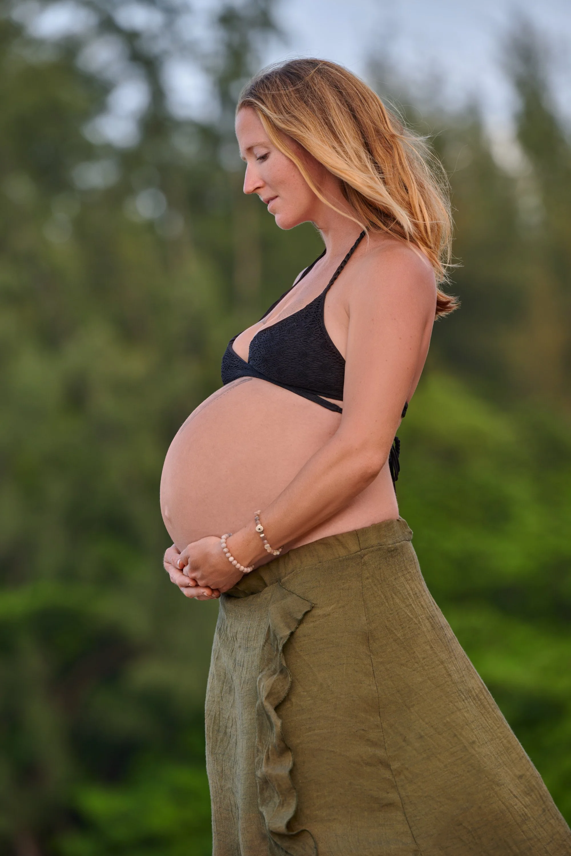 Profile in a black bikini top and olive skirt, jungle green rising behind.