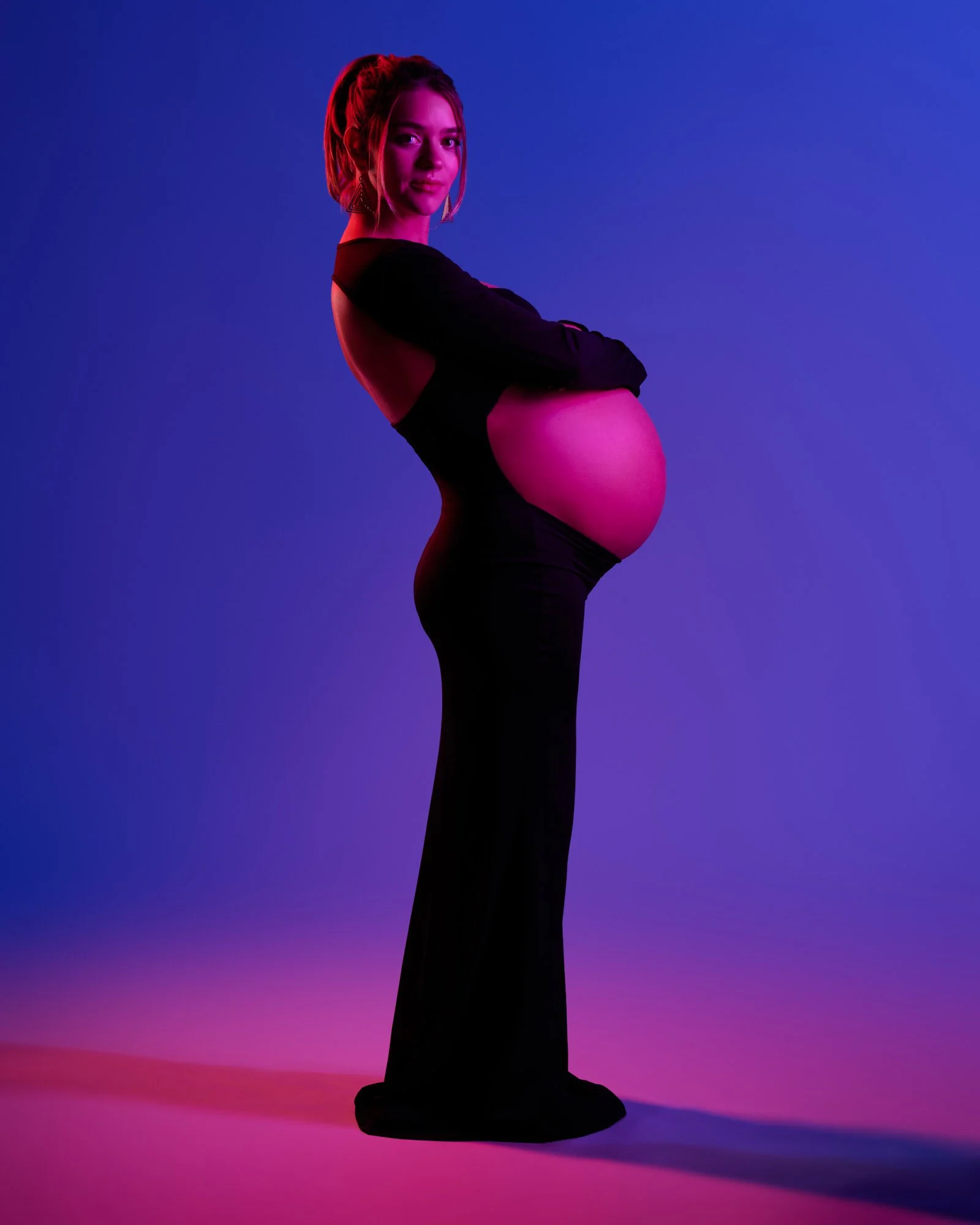 Studio profile in a long black gown with the bare bump lit against a magenta and blue backdrop.