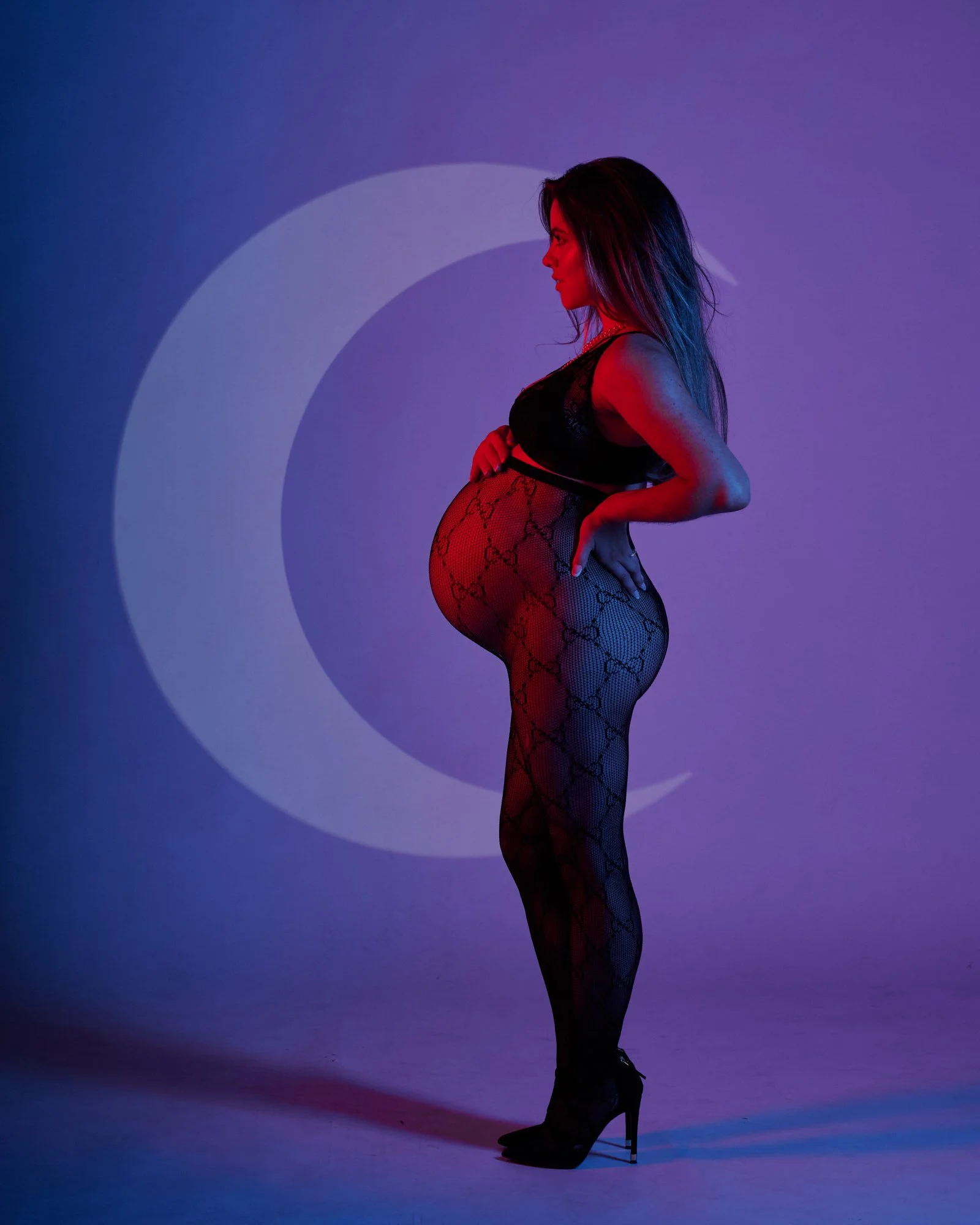A pregnant woman in black lingerie and patterned fishnet stockings standing in profile against a purple and blue background with a large crescent moon graphic.