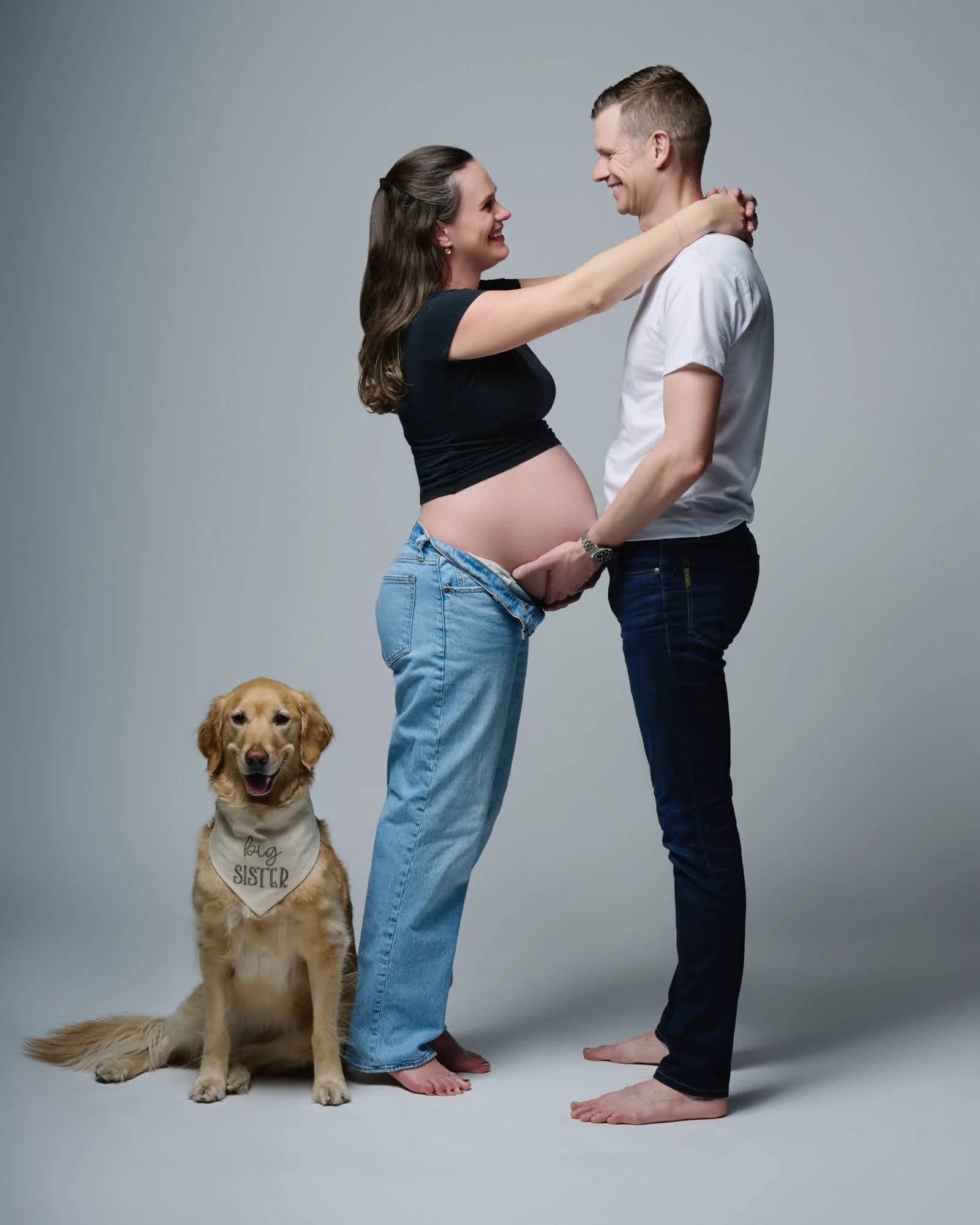 Couple laughs with their golden retriever in a big sister bandana during their studio session.