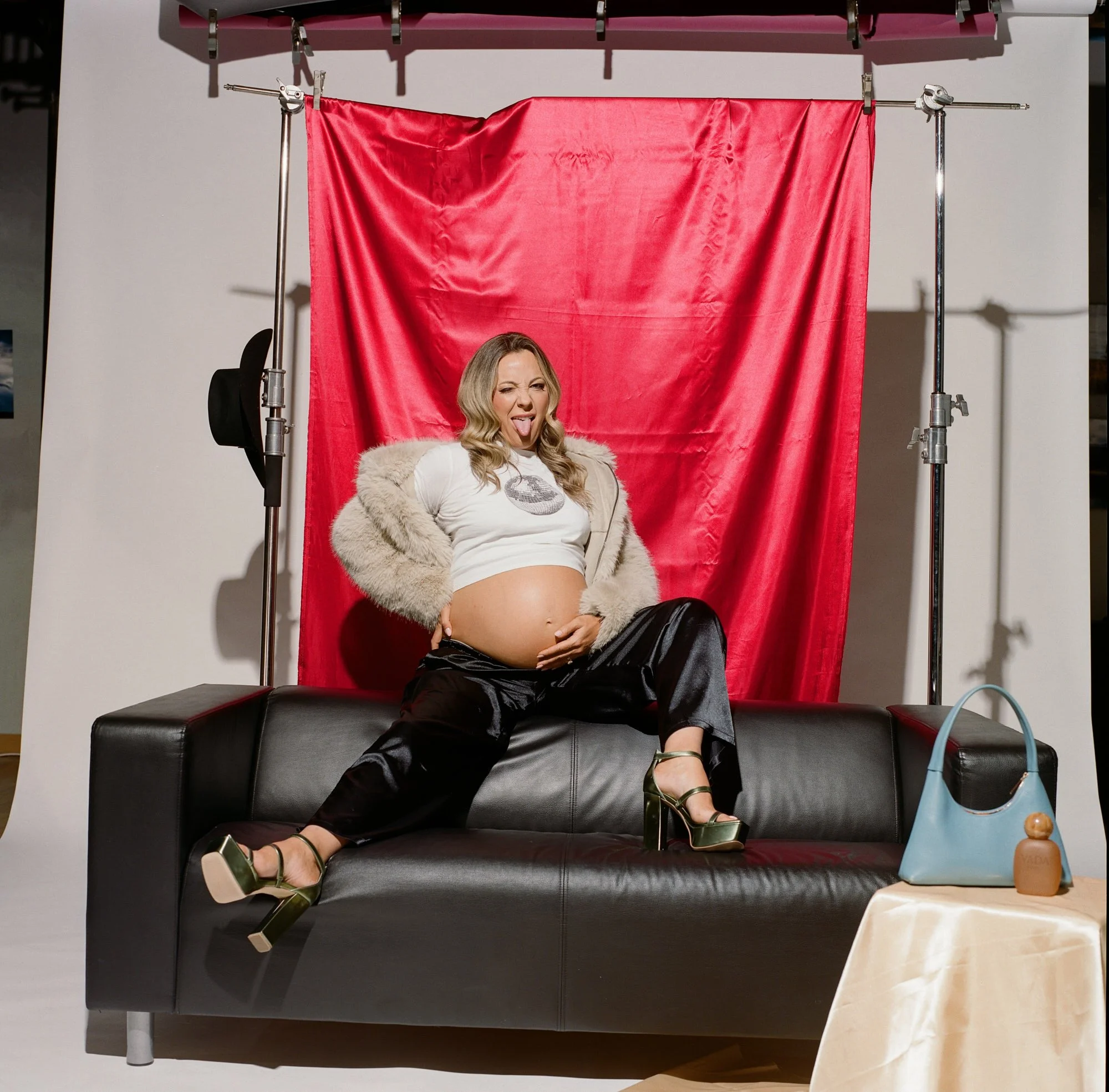 White fur coat and leather trousers on a black sofa, red backdrop lit and rigged behind her.