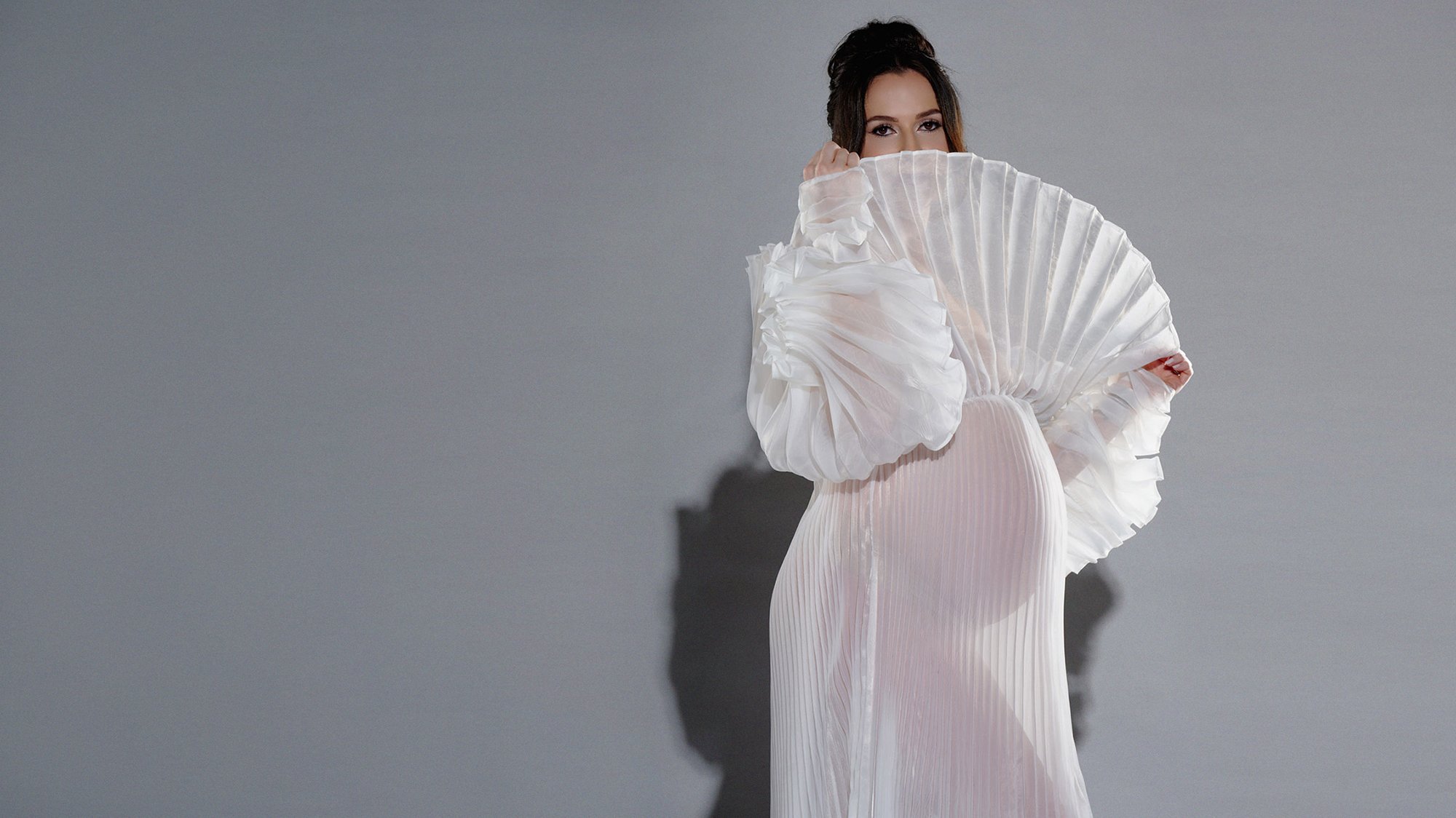 Expecting mother in a white pleated gown with fan sleeves against grey, one hand raised to her face.
