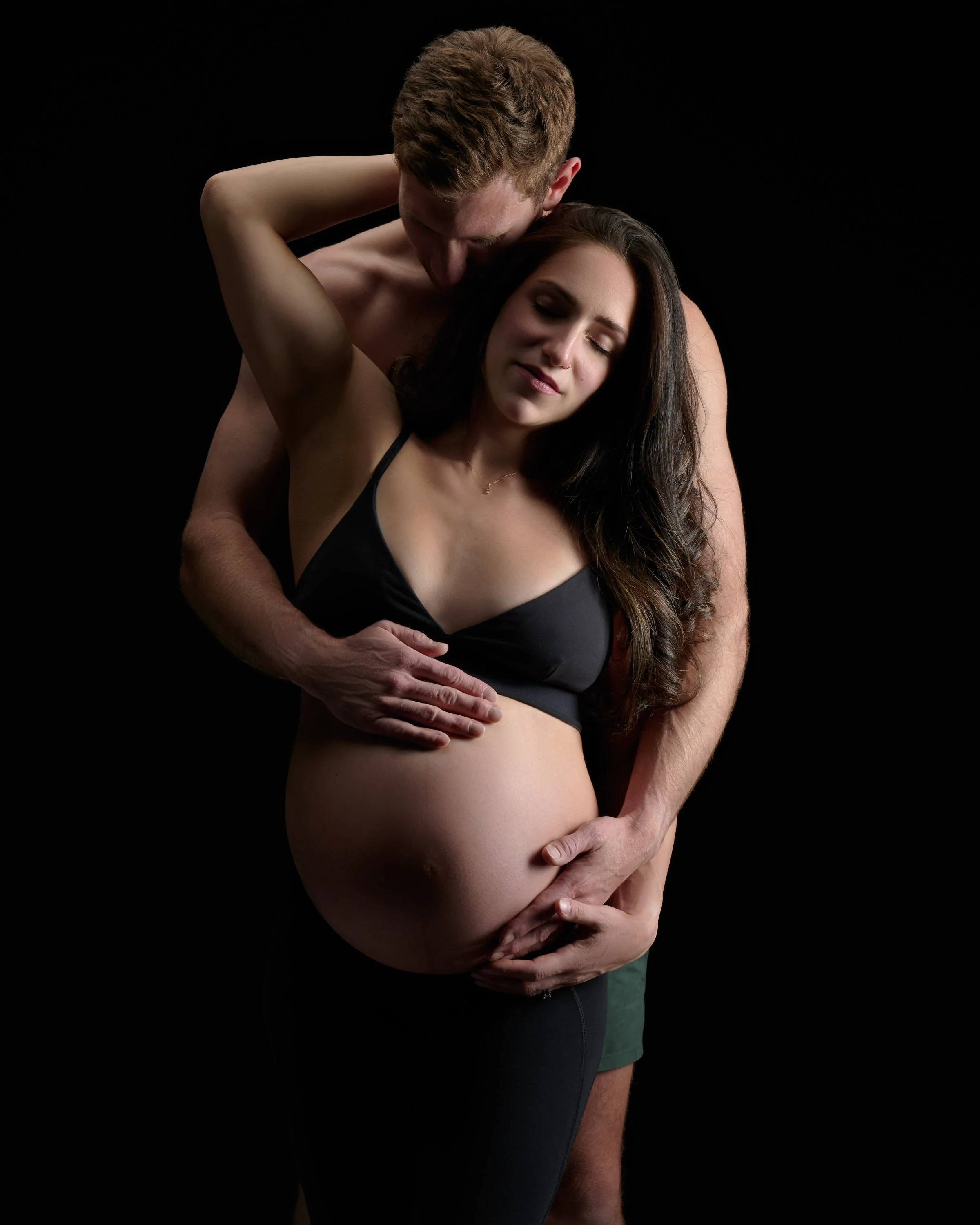 Partner kisses her head from behind, both hands resting on the bump against black.