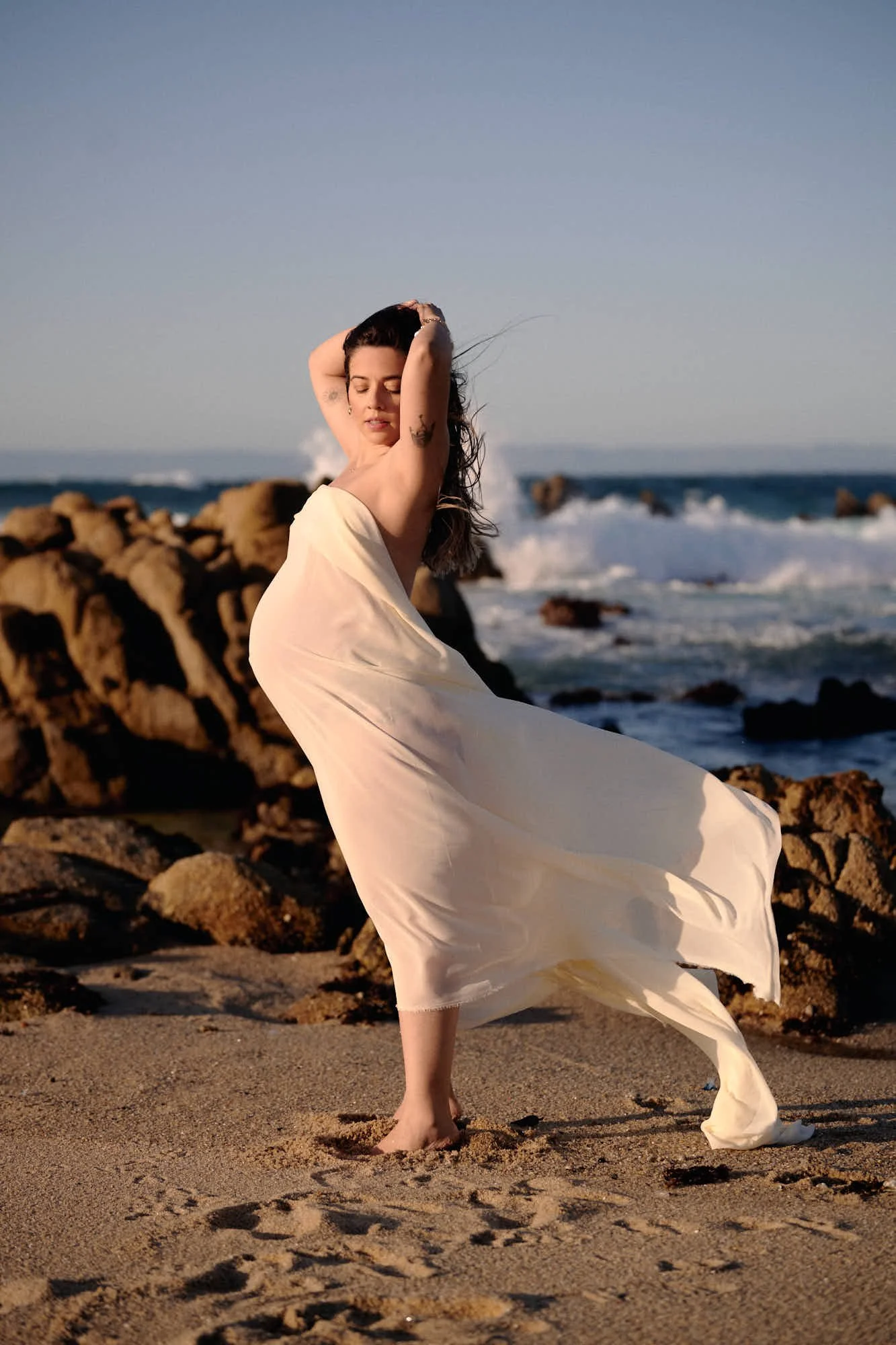 Sheer cream fabric lifting in the wind at the tide line, waves breaking over dark rocks.
