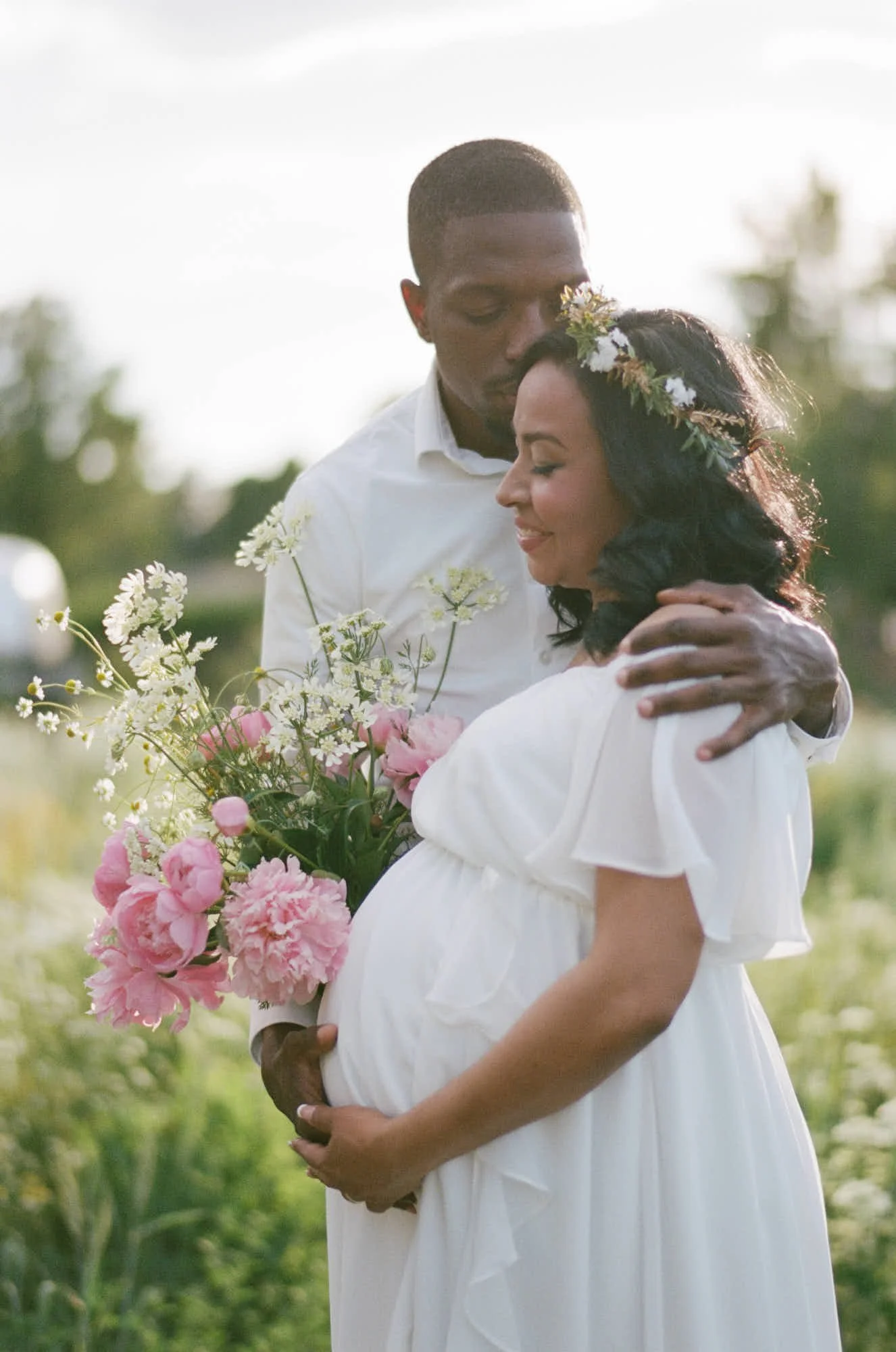 Expecting couple embracing in a garden near Lakewood, Colorado, mother in a flower crown with a pink bouquet, on film.