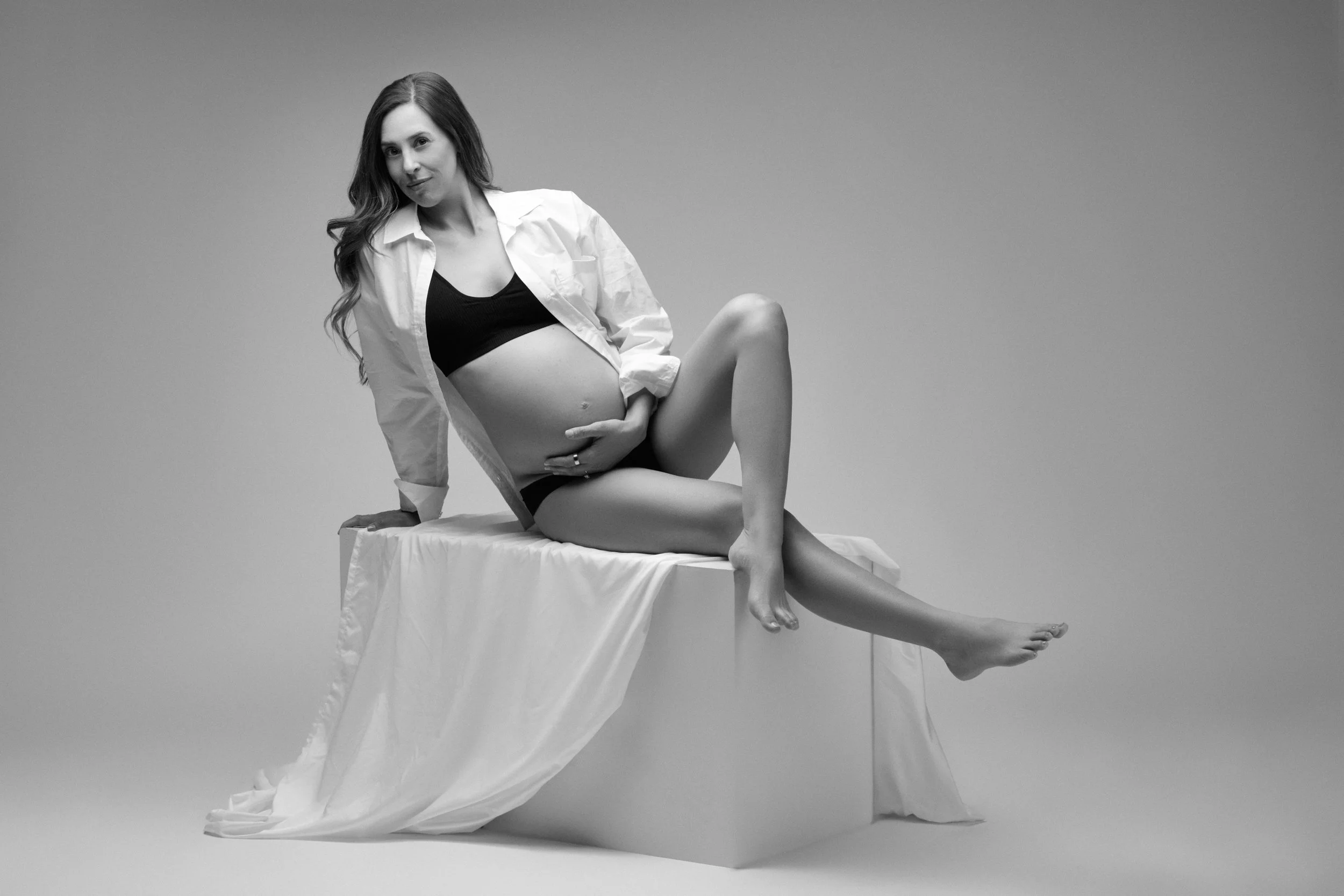 Black and white photo of a woman with long hair, sitting on a fabric-covered platform, wearing a white shirt and black undergarments, gently holding her pregnant belly.