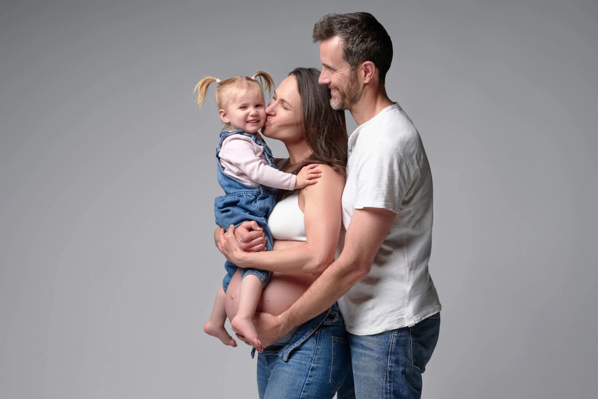 Mom kisses her giggling toddler while her partner wraps them both, a joyful family maternity portrait.