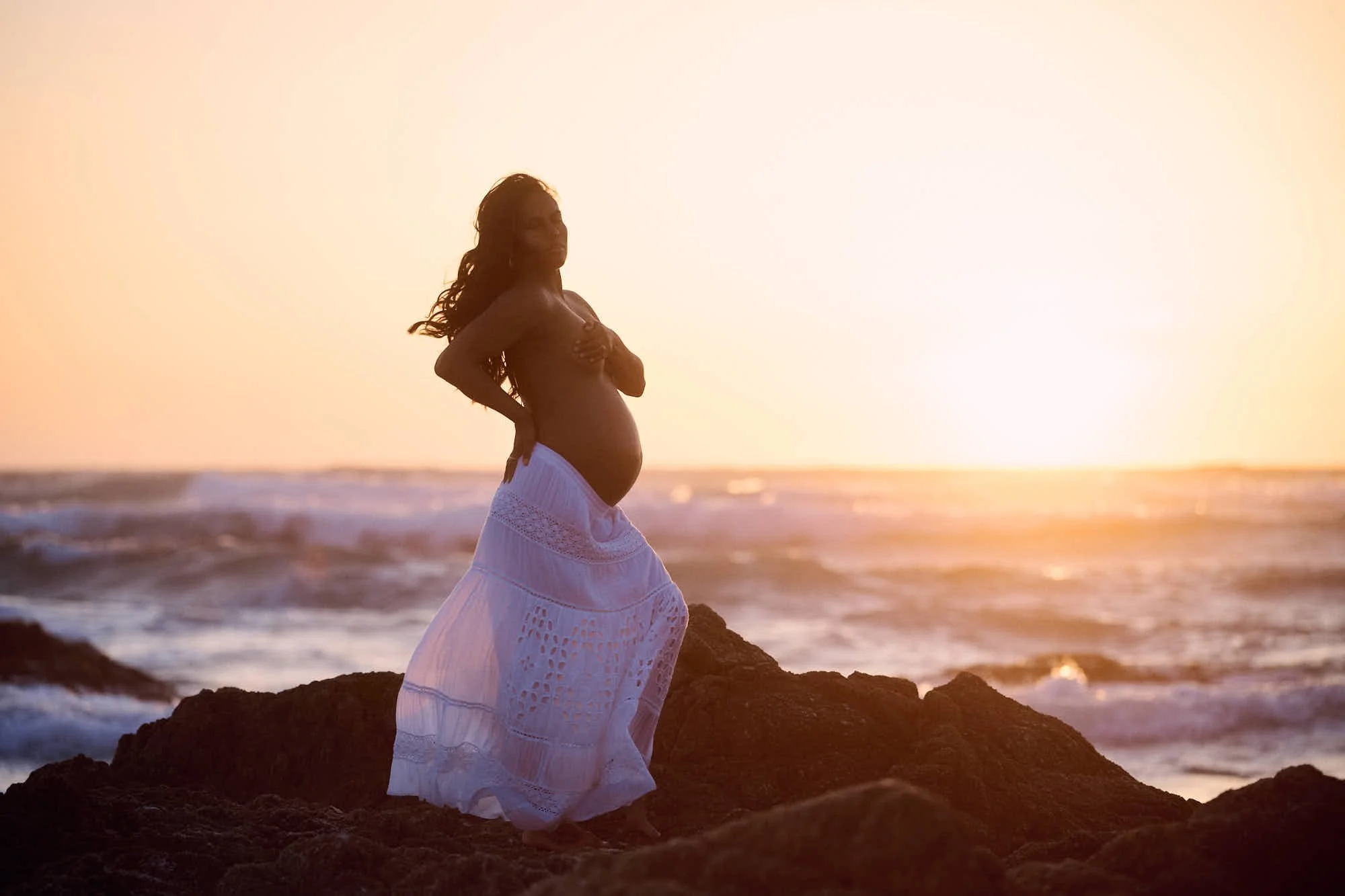 White dress on dark shoreline rocks, sun dropping into the Pacific behind the breaking waves.