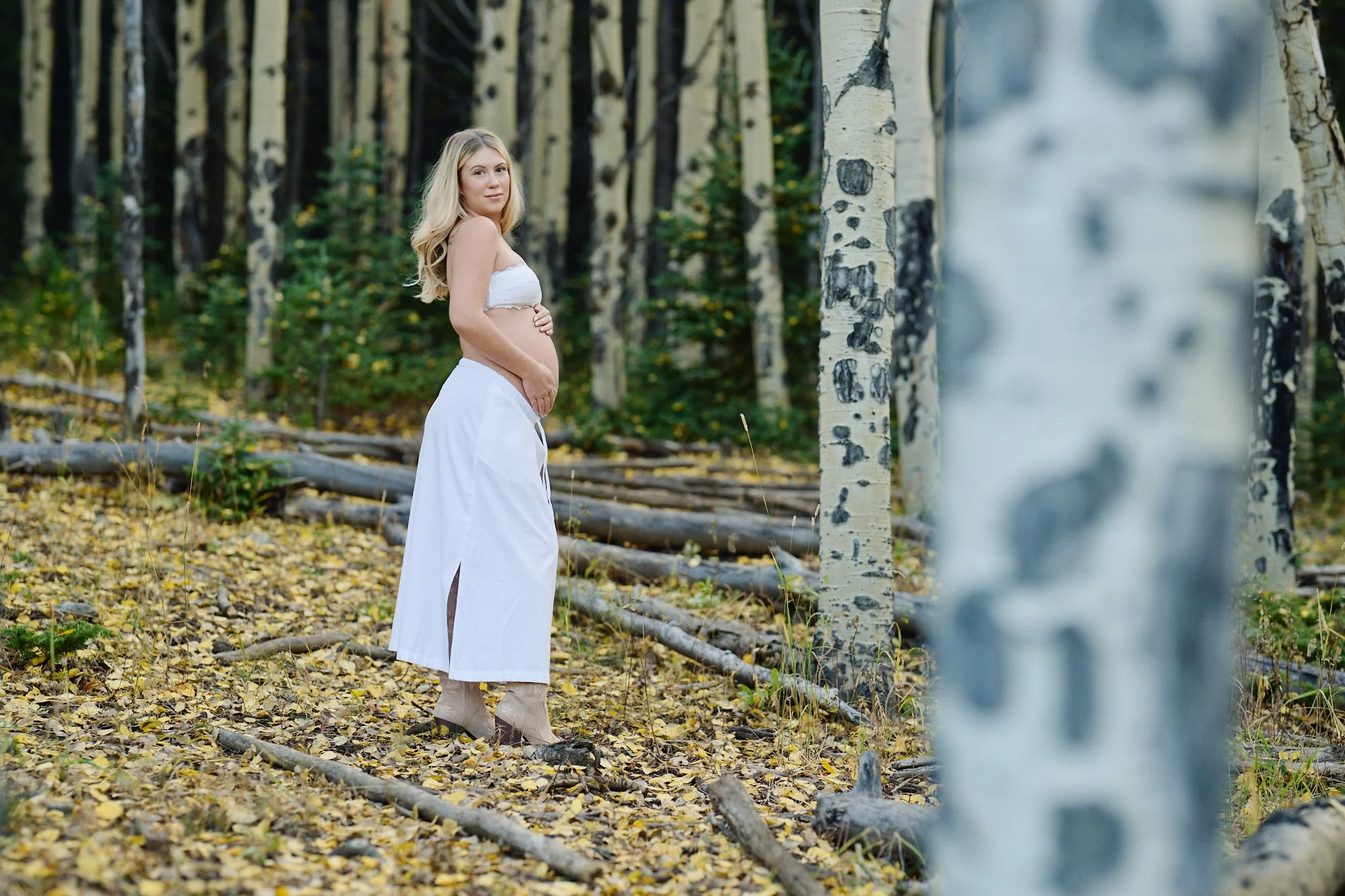 Mother-to-be in a white skirt standing in profile among aspen trunks, yellow leaves covering the forest floor.