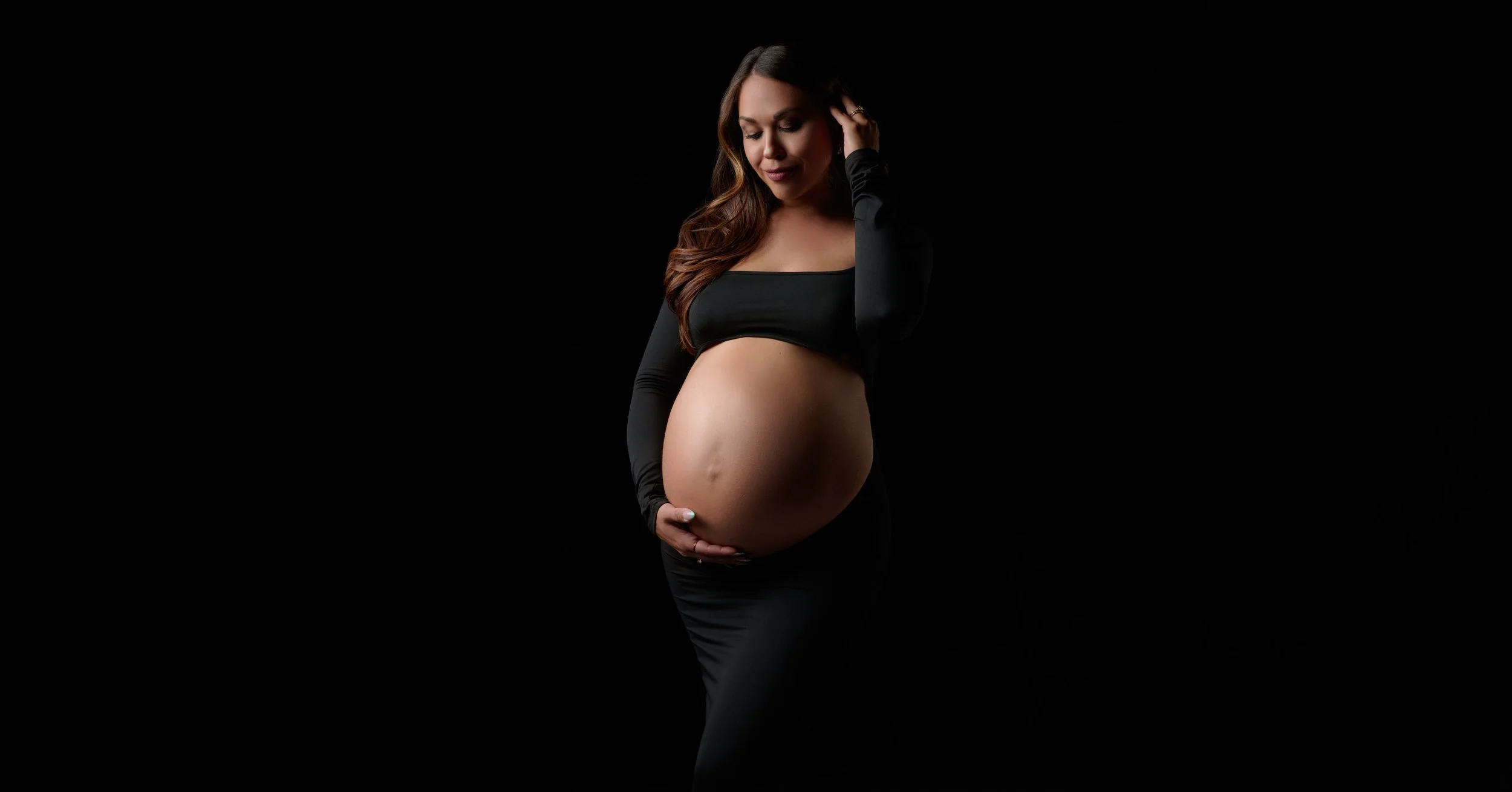 Black crop top above a bare bump, sculpted in moody light in our maternity photography studio in Denver.