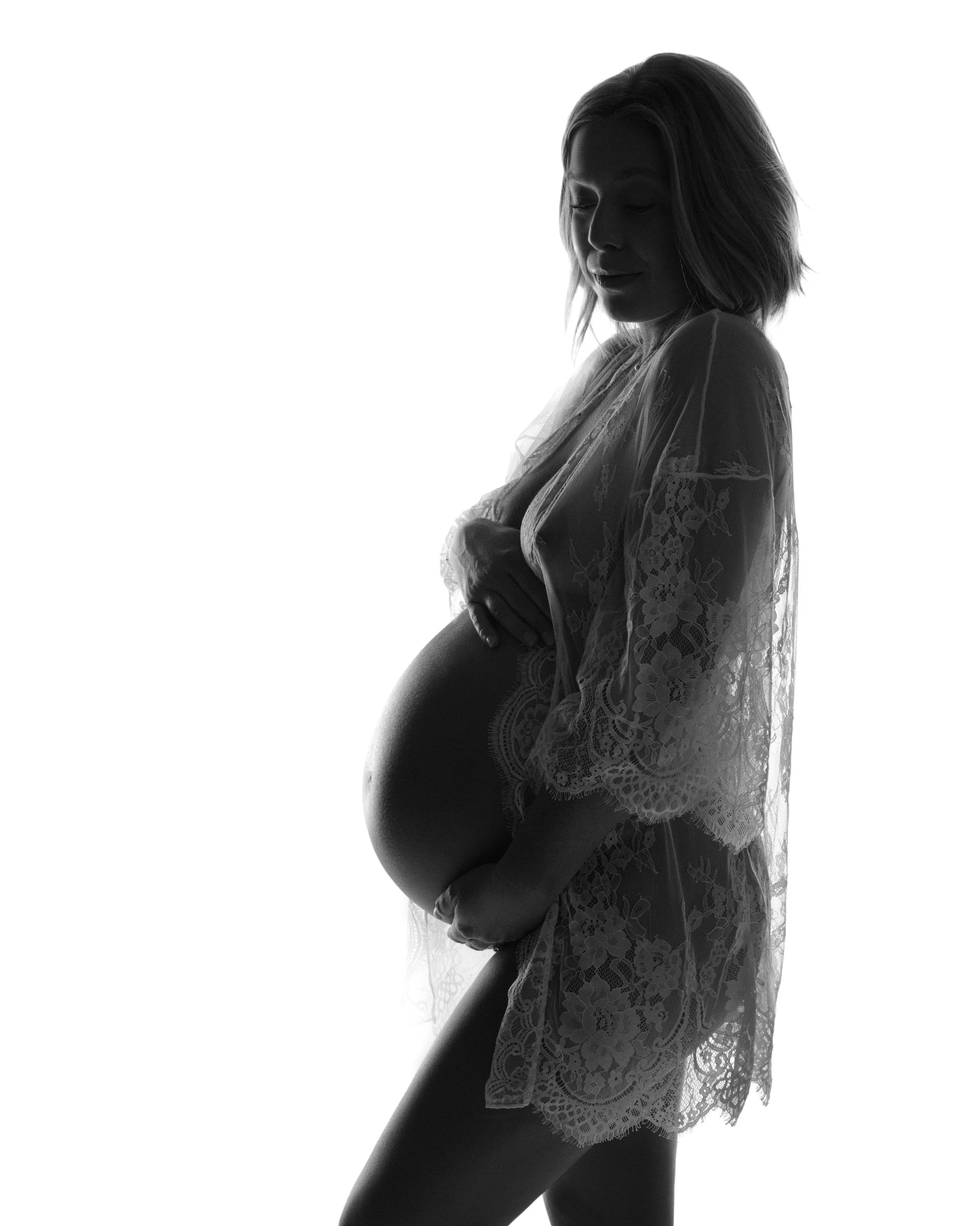 A sheer robe glows in a backlit silhouette, hands framing the bump.