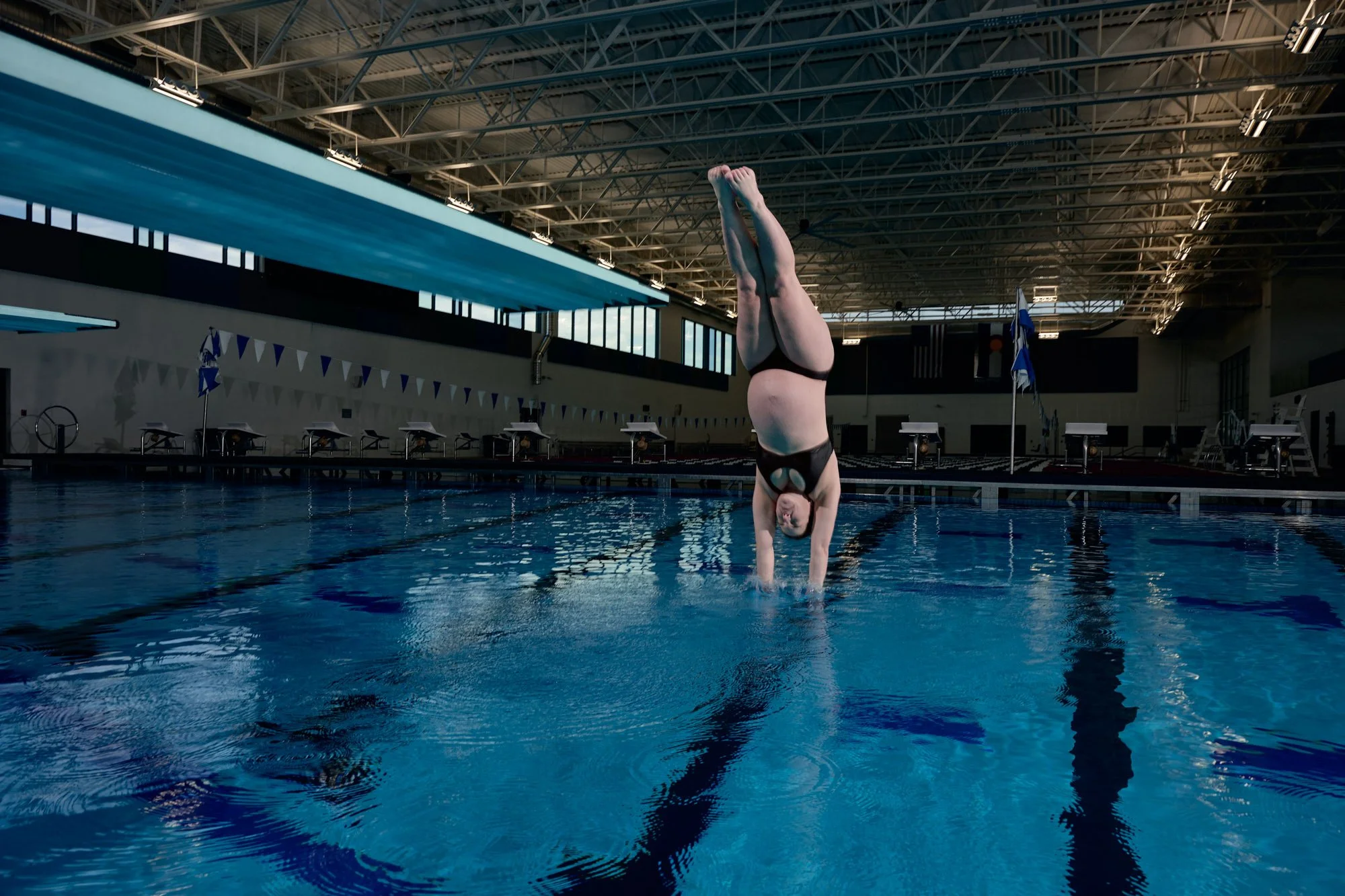 Wide natatorium frame with a diver inverted in a handstand at the far platform.