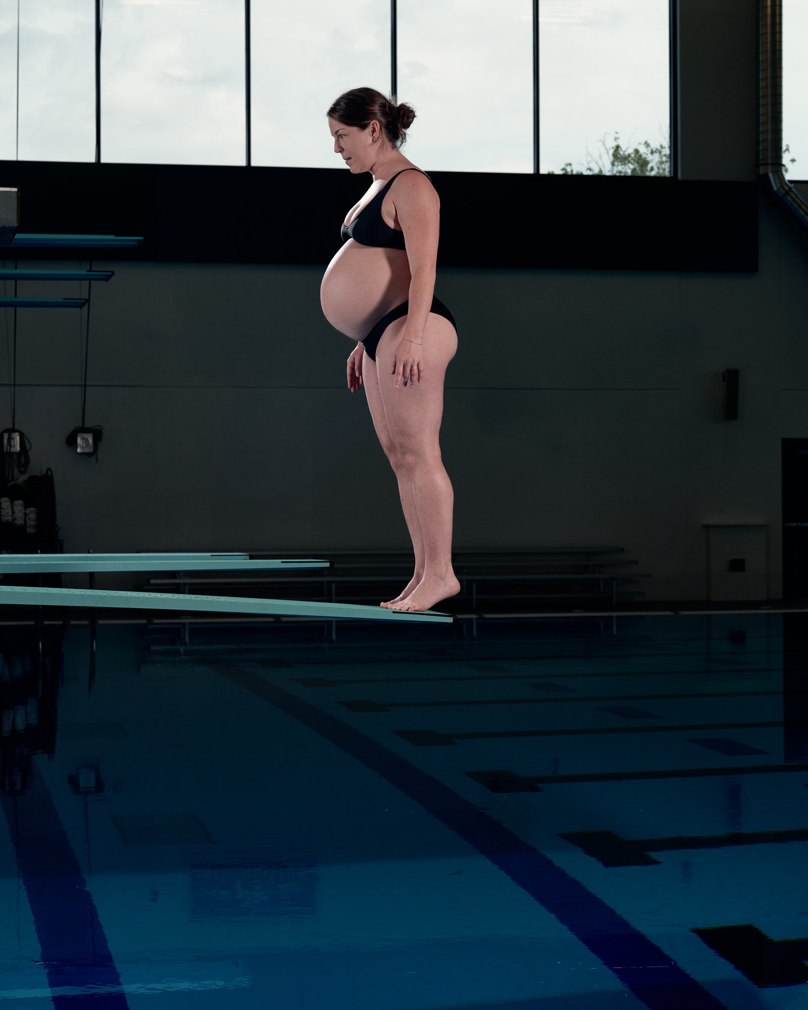 Profile at the edge of the board, hands behind her back, dark natatorium windows beyond.