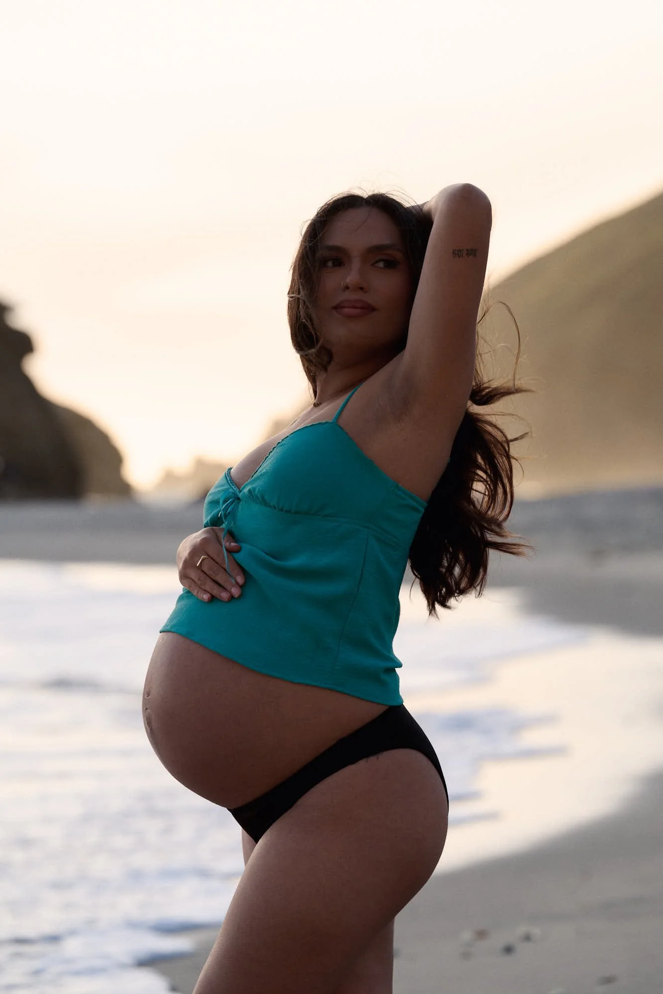 Teal top and bare bump on the sand at dusk, headland and surf soft in the background.