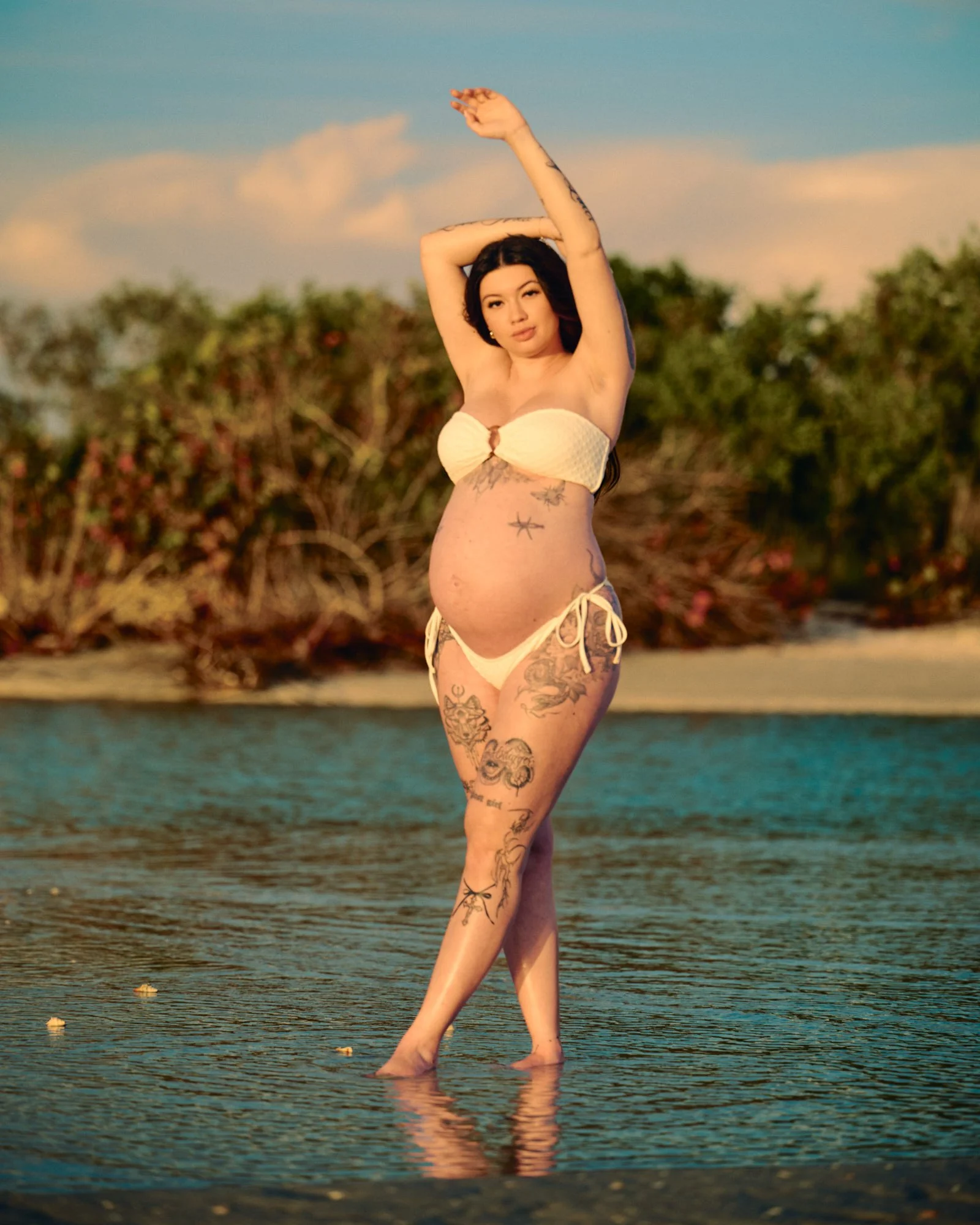 One arm raised in a white two-piece at the water's edge, mangroves green behind.