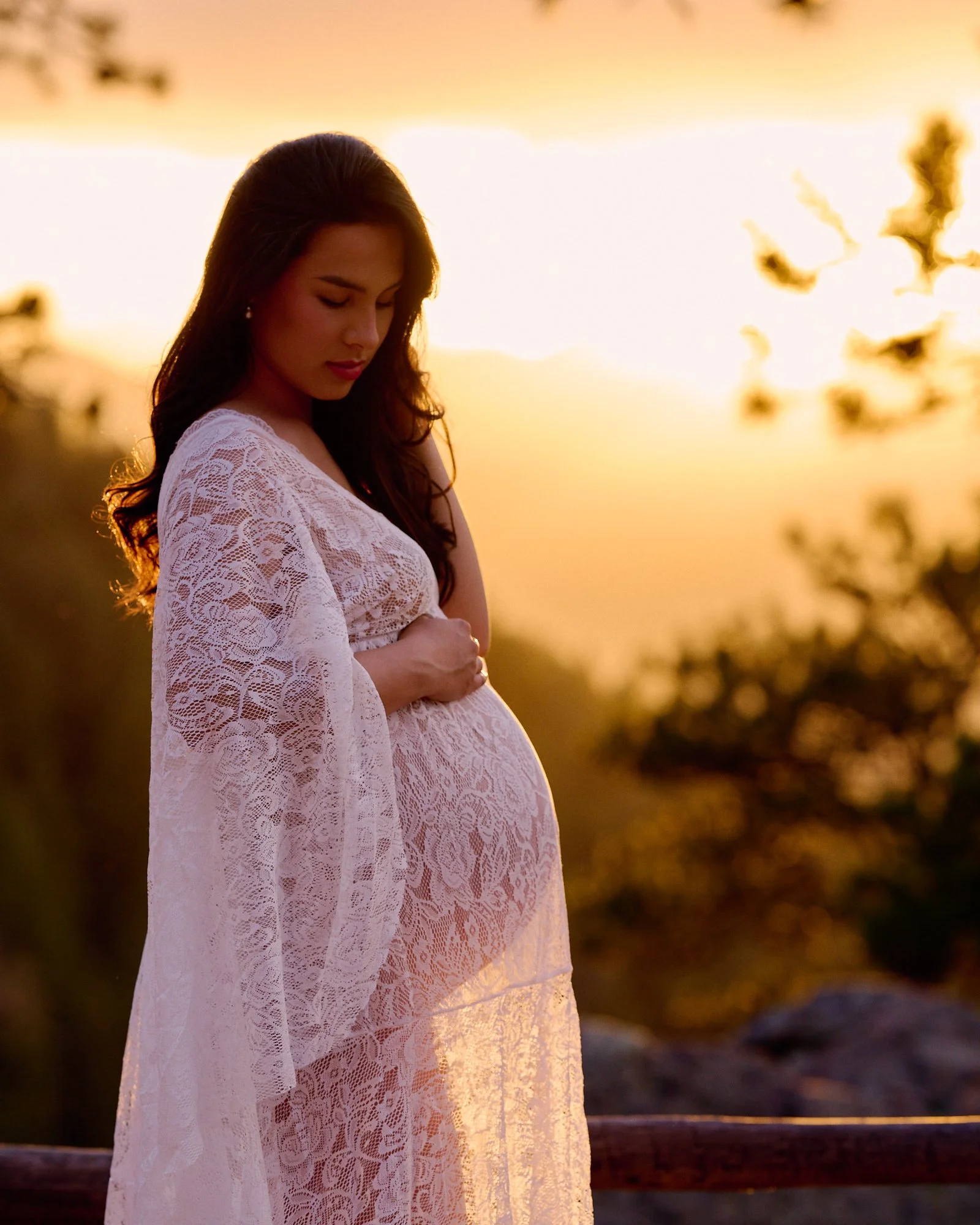 Golden hour glow through a white lace duster as she looks down at her bump.
