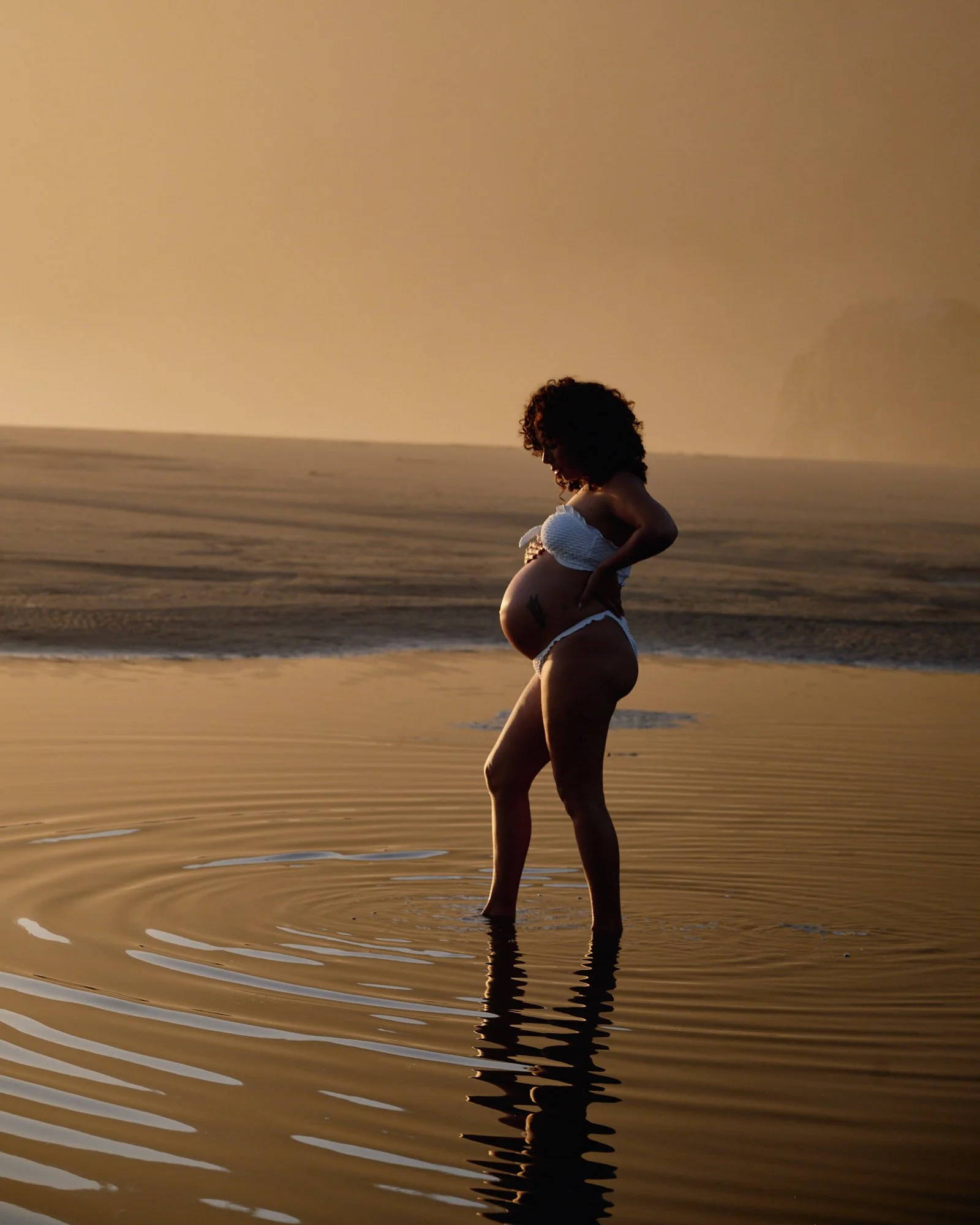 White two-piece in ankle-deep water at golden hour, ripples spreading from her feet across the flats.