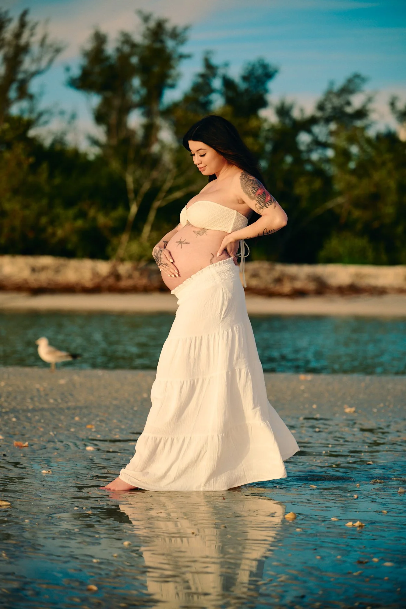 Wading through shallow water in a white tiered skirt, a shorebird on the sandbar beside her.