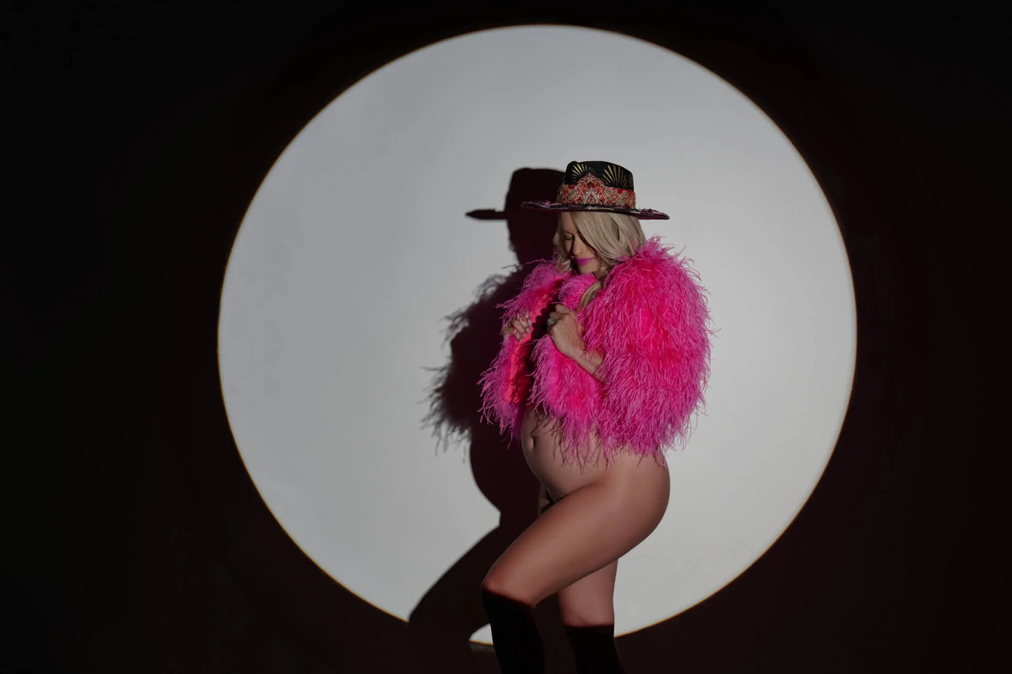 Wide-brim hat and pink feather jacket inside a spotlight circle, pure editorial attitude.