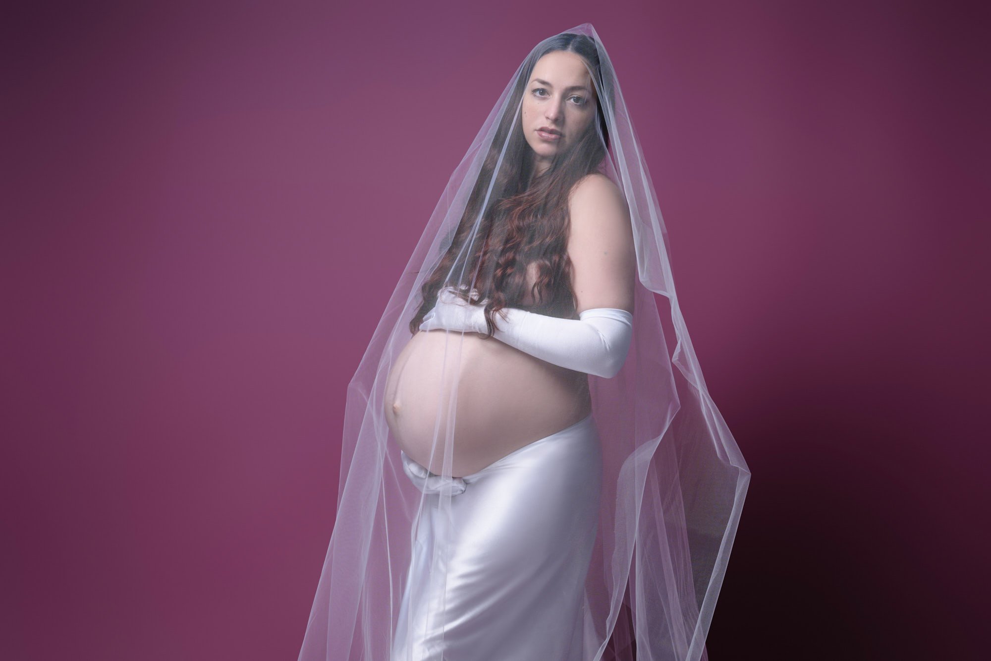Satin skirt, opera gloves, and a long veil against a plum backdrop, arms crossed under her bump.