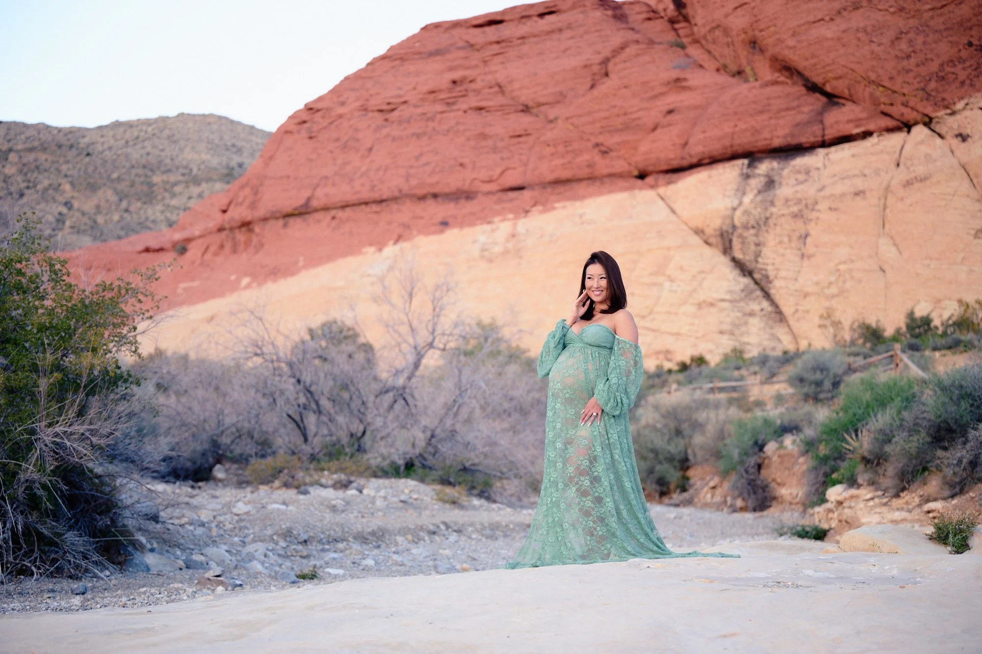 Small in the frame beneath a red sandstone wall, sage lace gown in Red Rock Canyon.