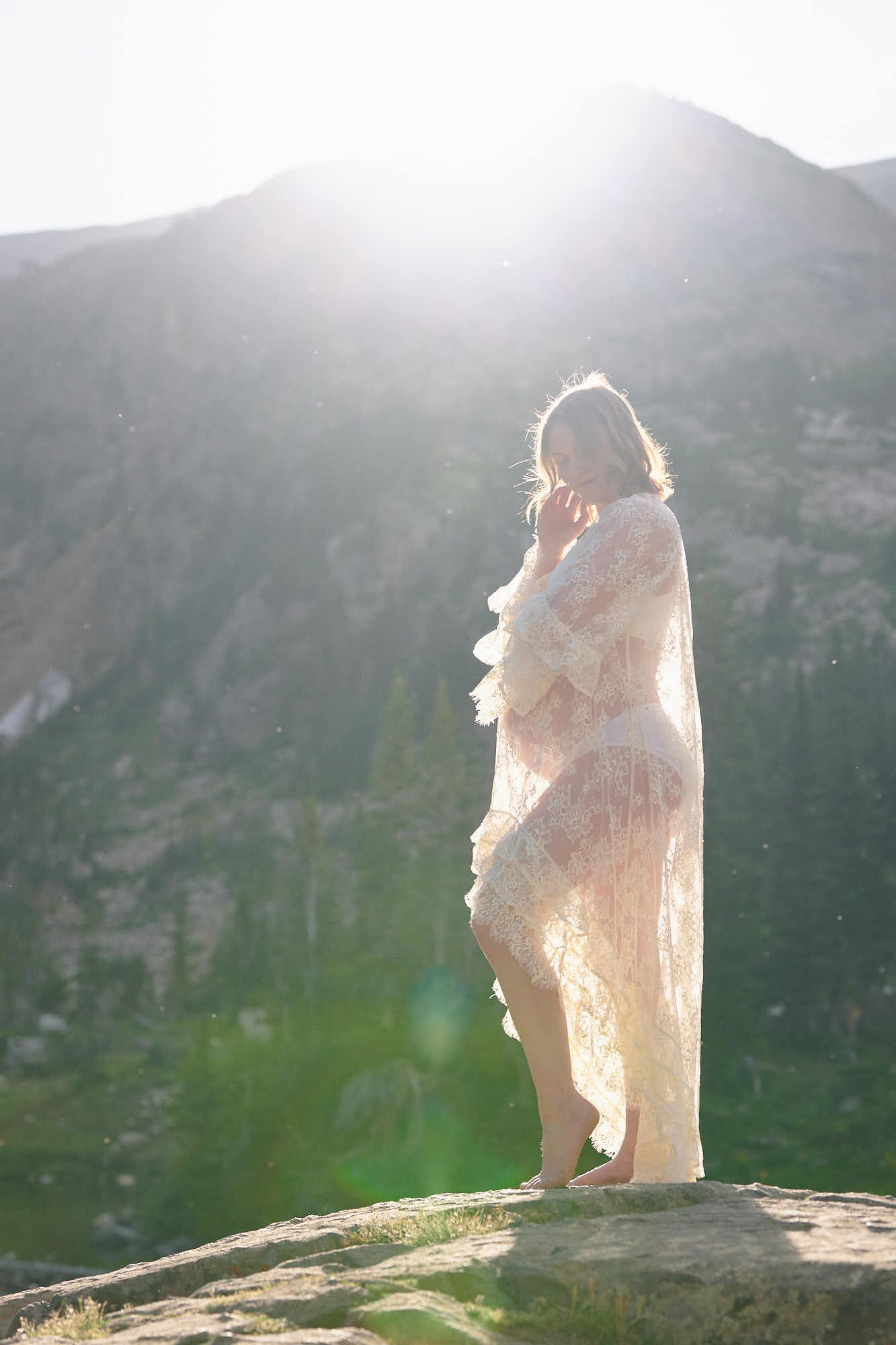 Expecting mother in a sheer ivory lace robe, backlit by sun flare on a Colorado alpine ridge.