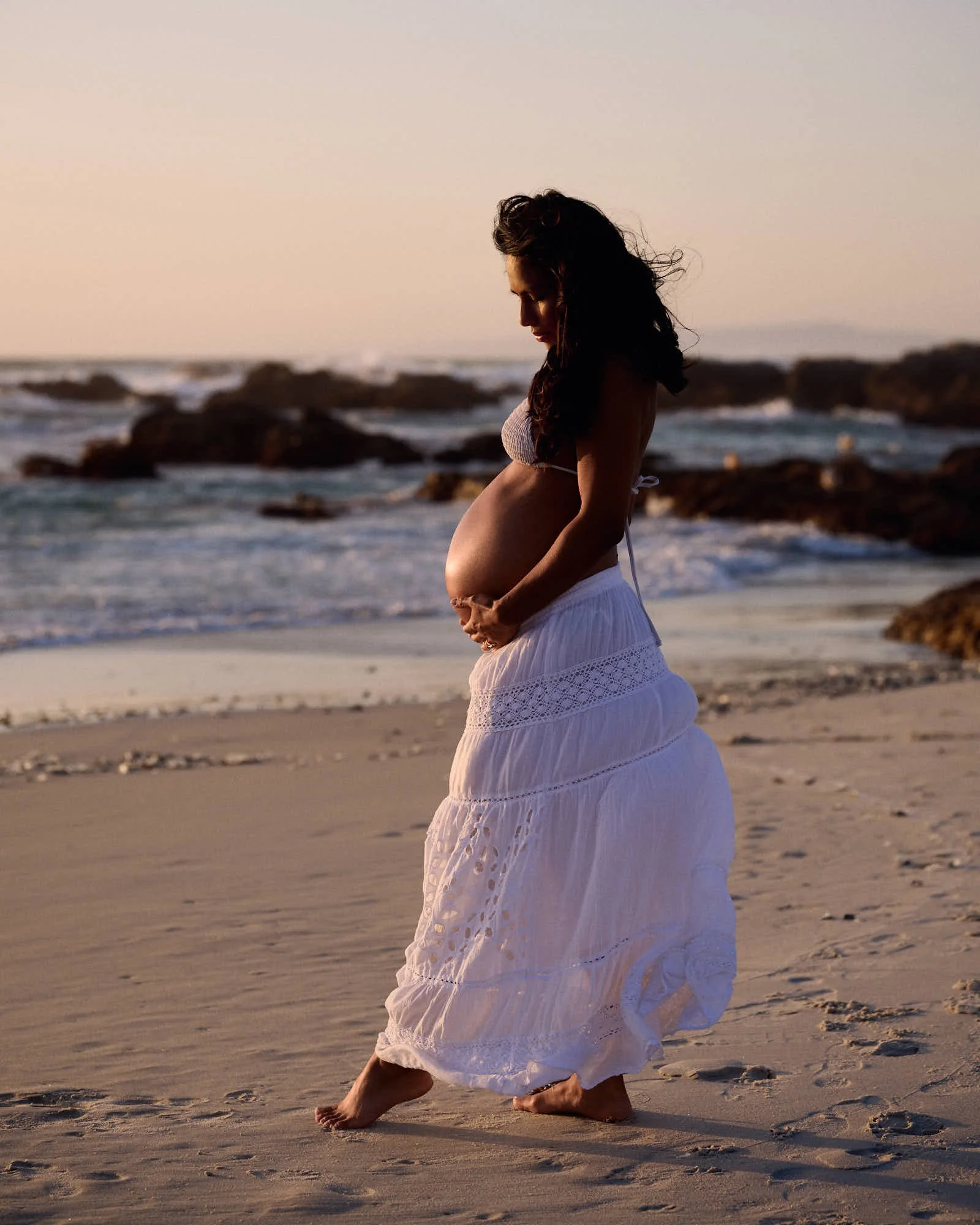 Walking the wet sand in a white lace skirt, hands on the bump, rocks and surf behind.