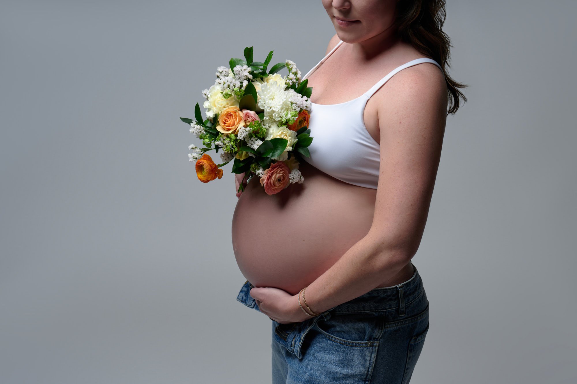 Peach bouquet held above a bare bump, white bra and denim, a soft close-up.