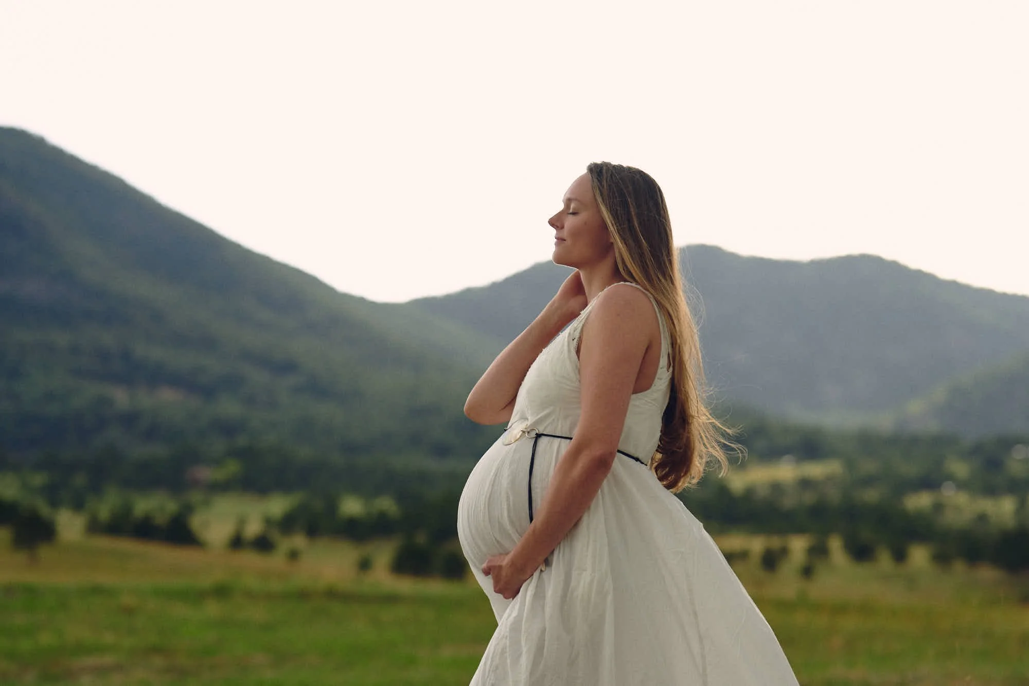 White sundress with navy ties in a deep green mountain bowl, hand at her collar.