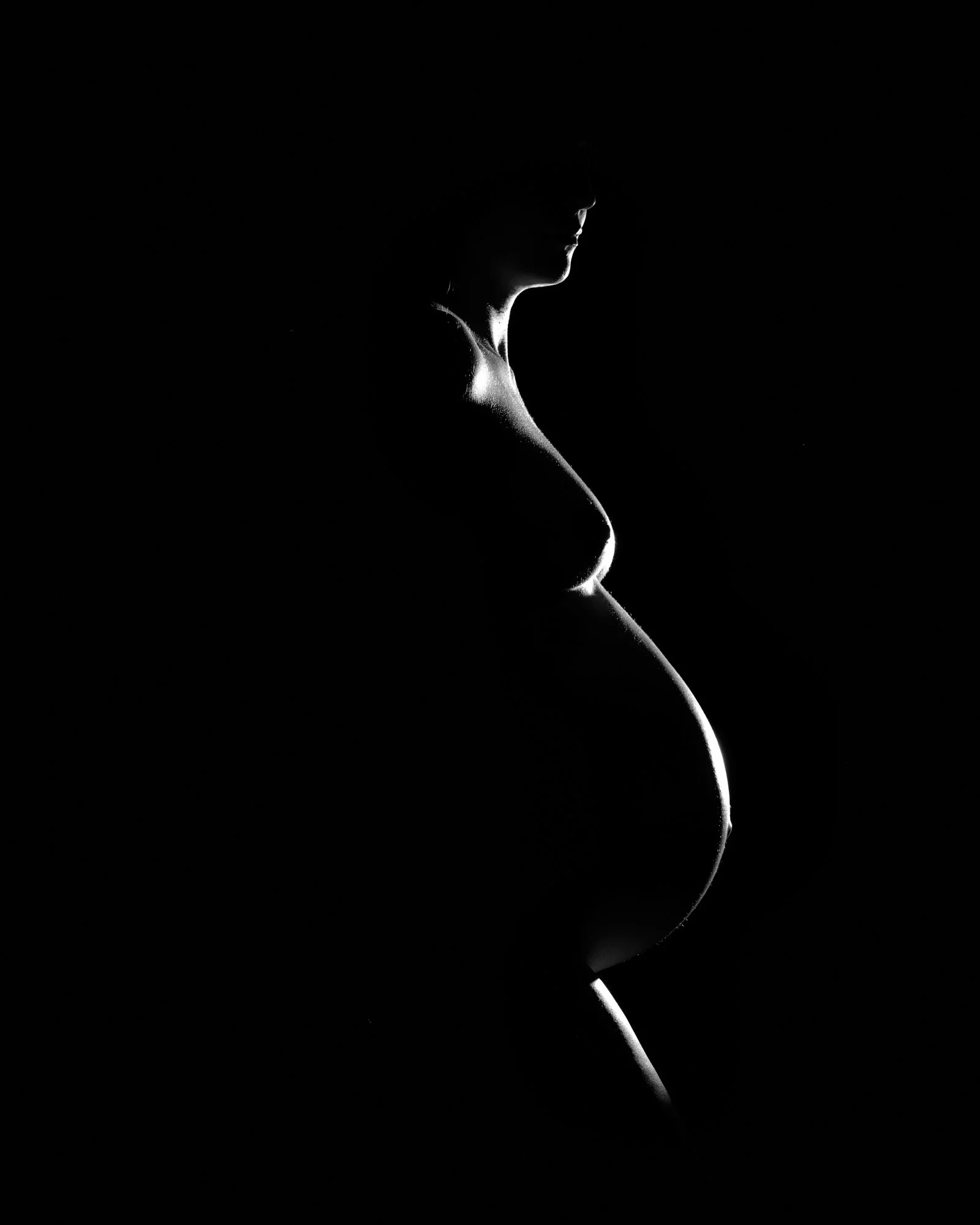 Rim light traces a pregnant profile and bump out of pure black, a fine art silhouette.