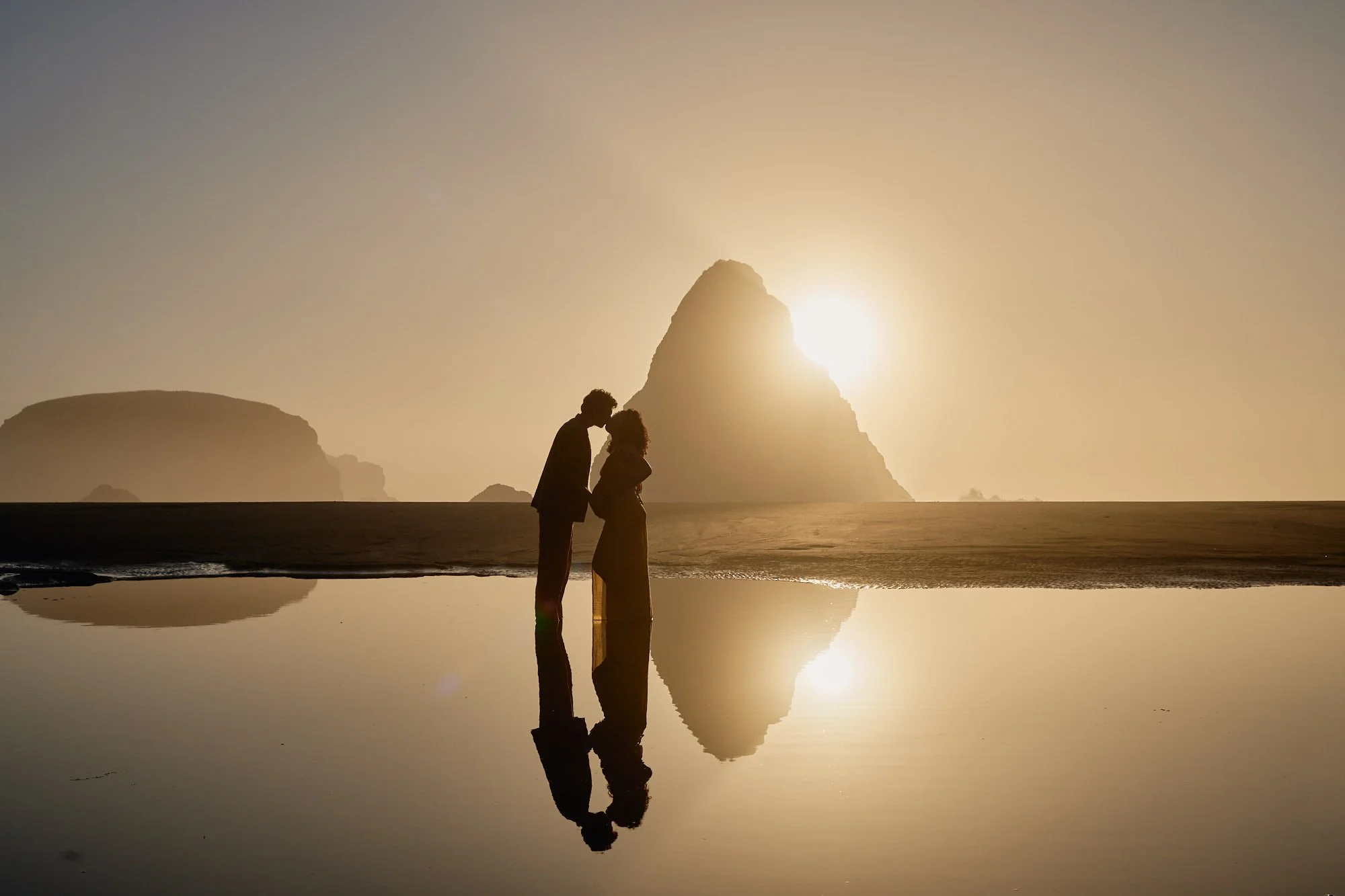 Couple kissing in silhouette on the tide flats, sun setting behind a sea stack and their reflection below.