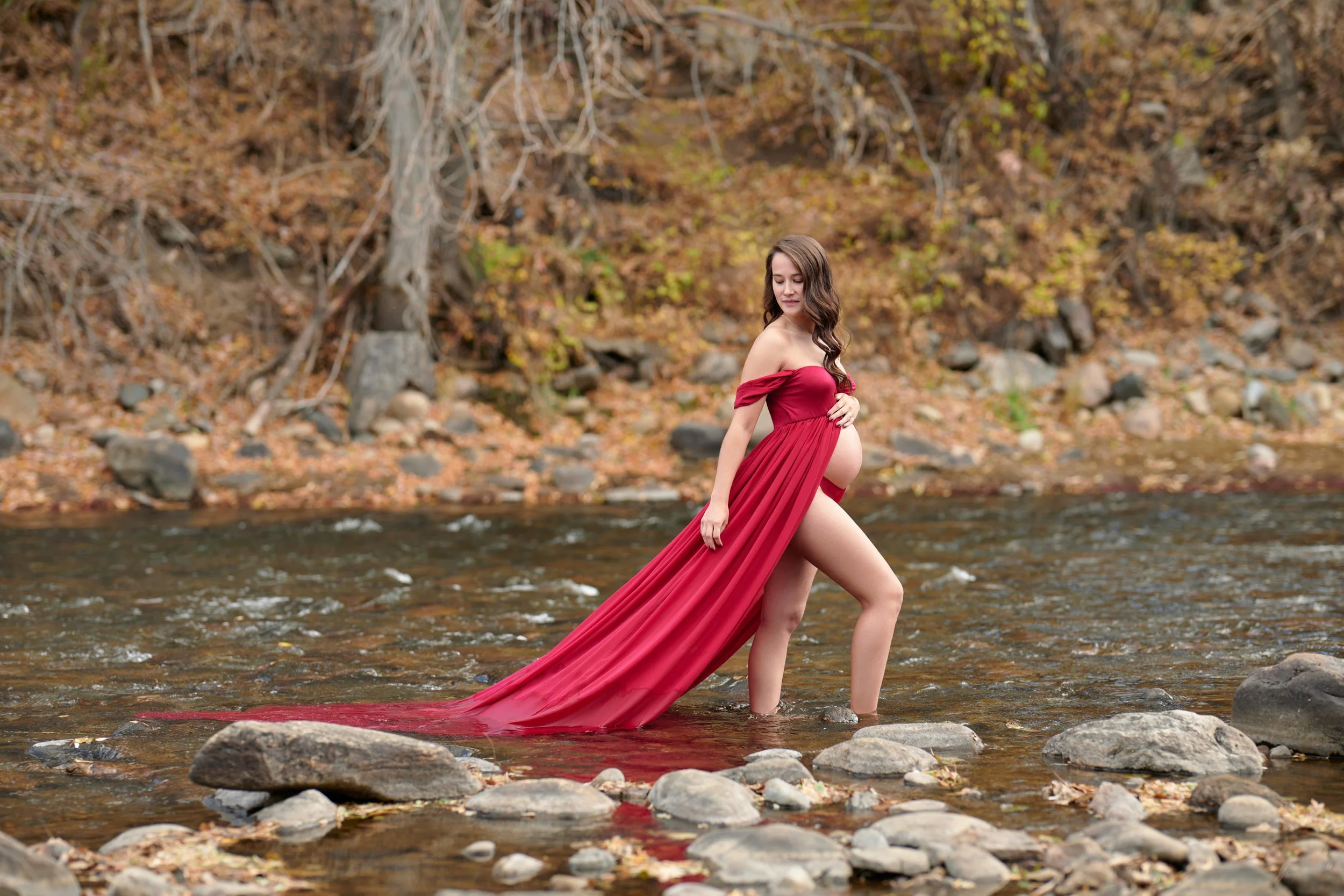 Crimson gown wading a rocky autumn creek, train trailing the current.