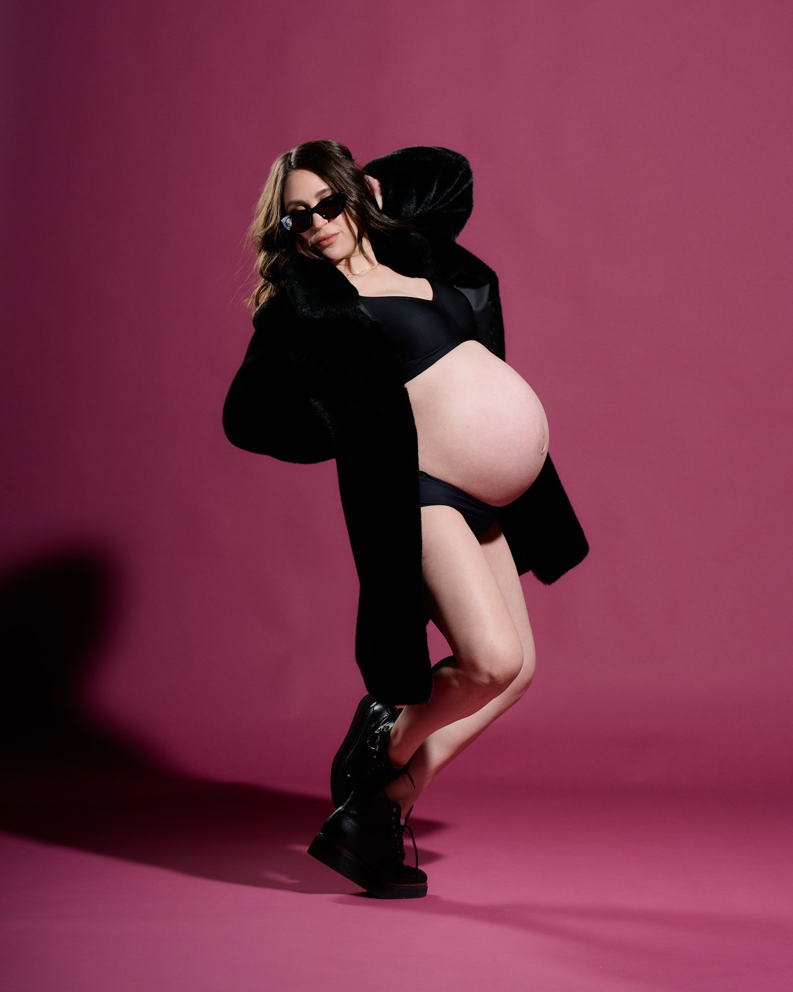 Expecting mama in a black fur coat and sunglasses striking a playful pose against a magenta backdrop.