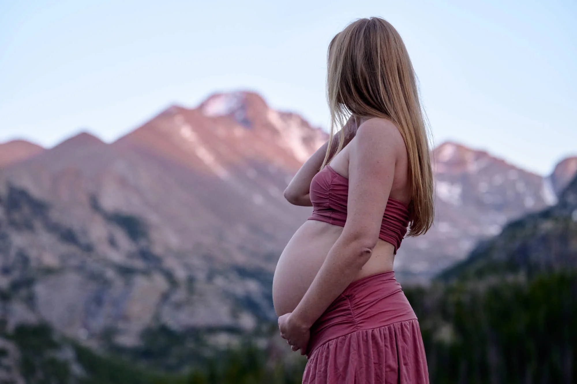 Expecting mother in dusty rose cradles her bump on an alpine overlook as alpenglow hits the peak.
