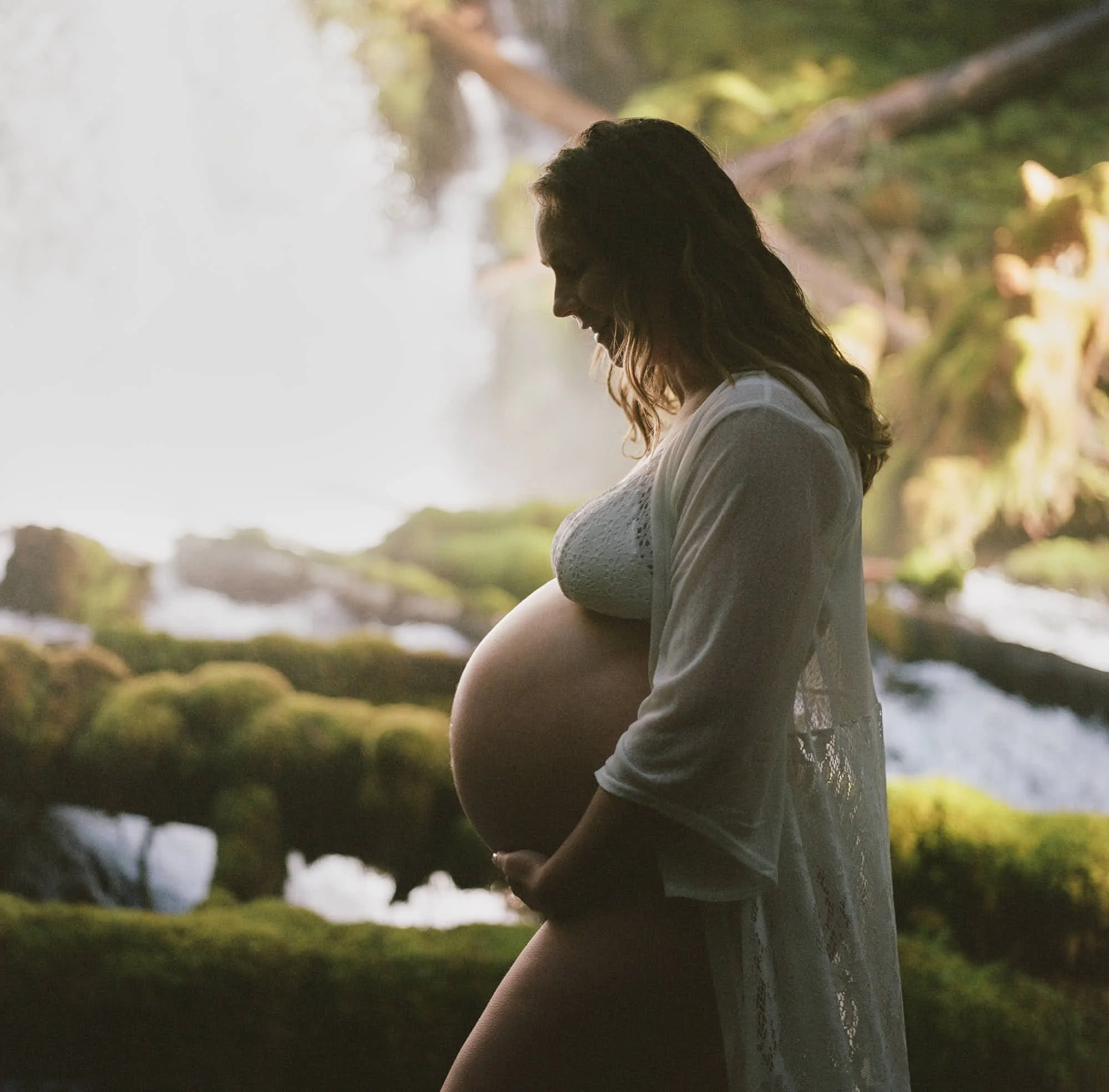 Profile in a white lace robe against the falls, backlight rimming the bump, shot on color film.
