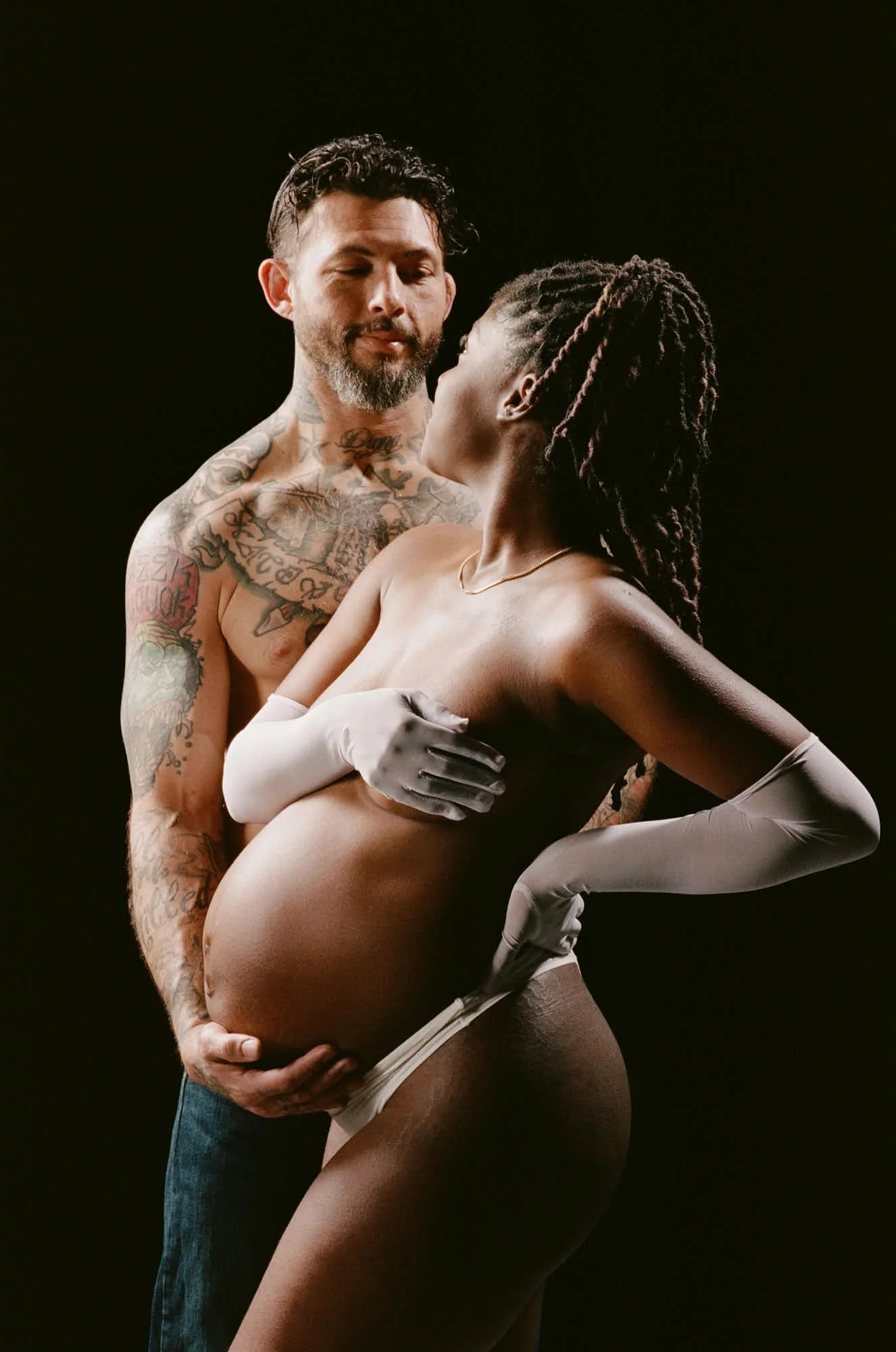 Partner standing behind a mother-to-be in long satin gloves, hands together on her bump in low light.
