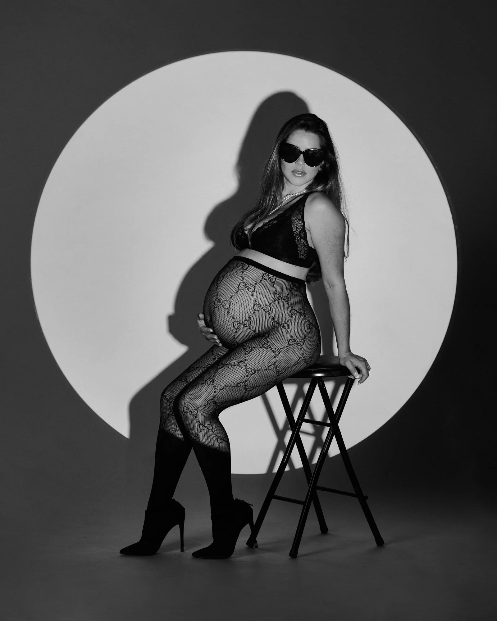 A woman wearing sunglasses, black lingerie, fishnet stockings, and high heels, posing on a stool with a large circle lighted background, creating a shadow on the wall.