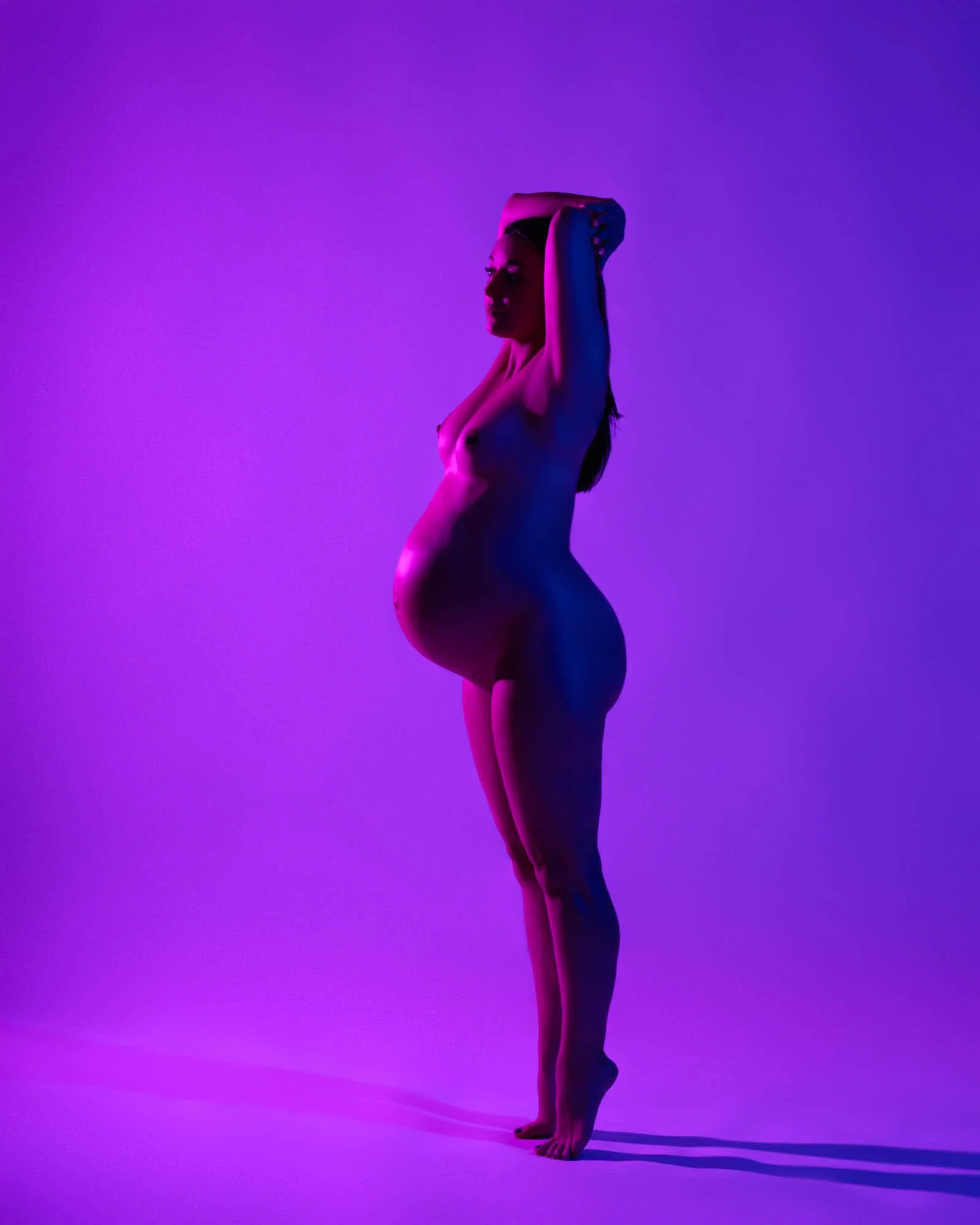 Statuesque on tiptoe under purple gel light, arm arched overhead.