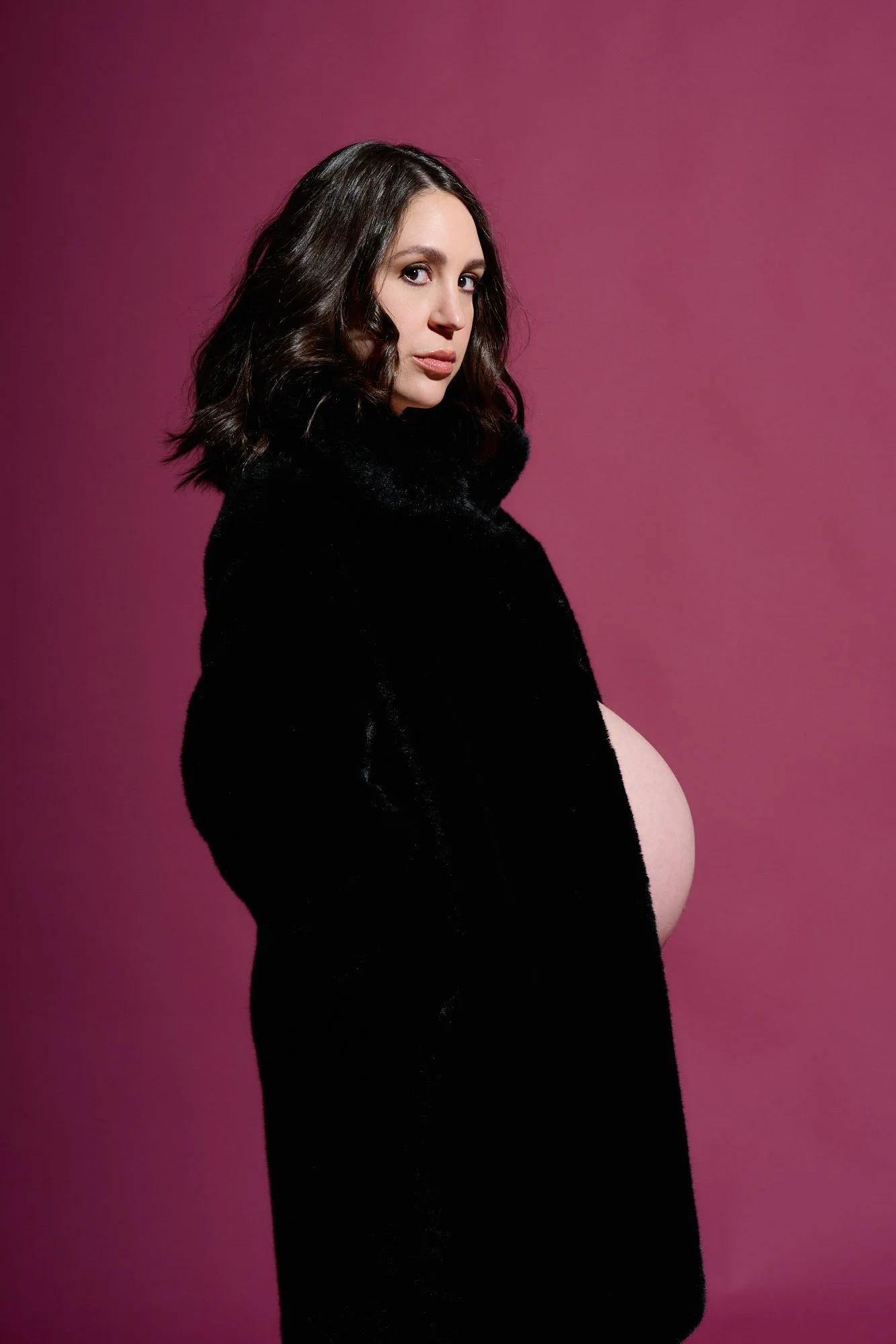 Black coat open over a bare bump against a deep berry backdrop, editorial and still.