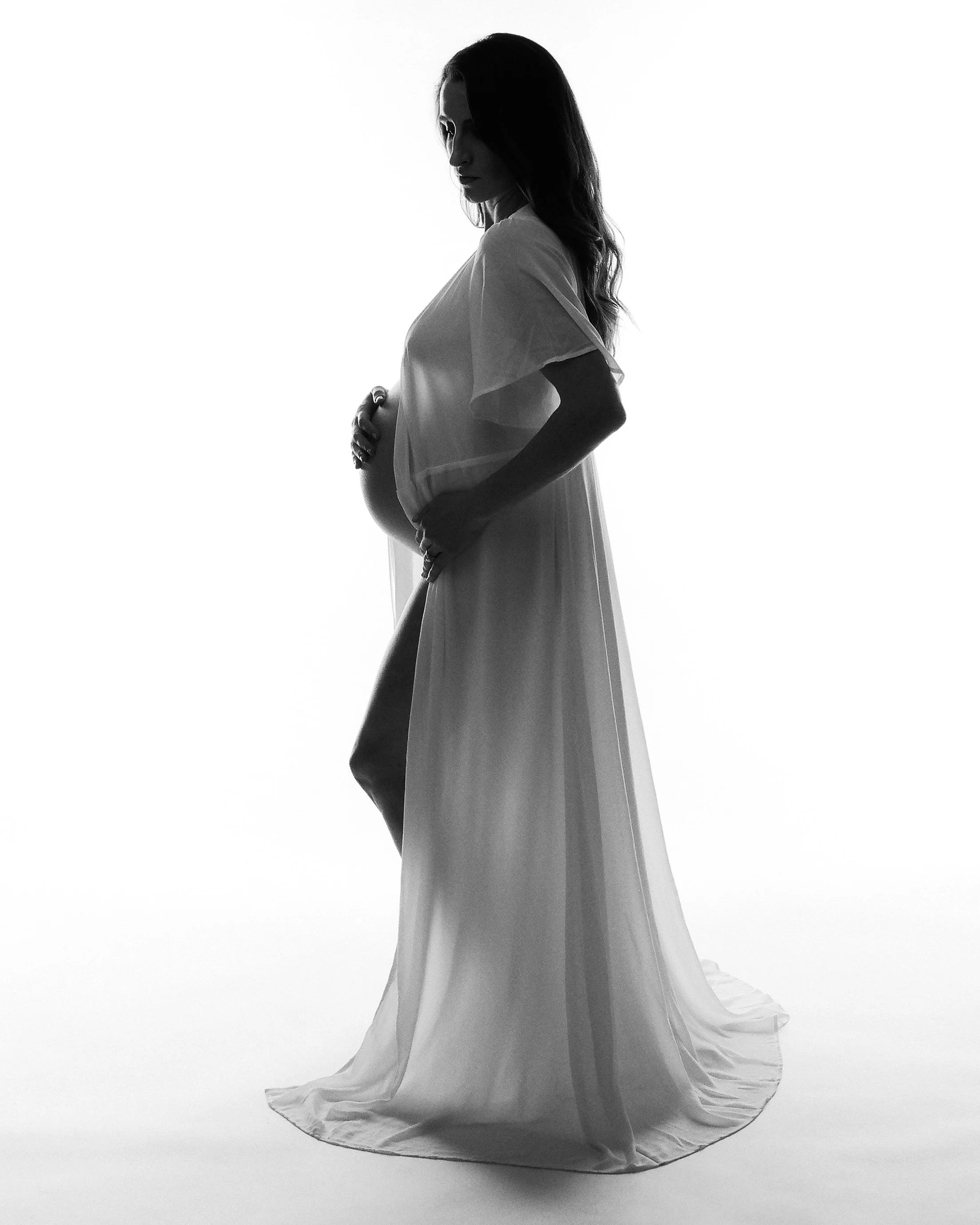 Sheer gown caught mid-motion, fine art maternity portrait in high key black and white.
