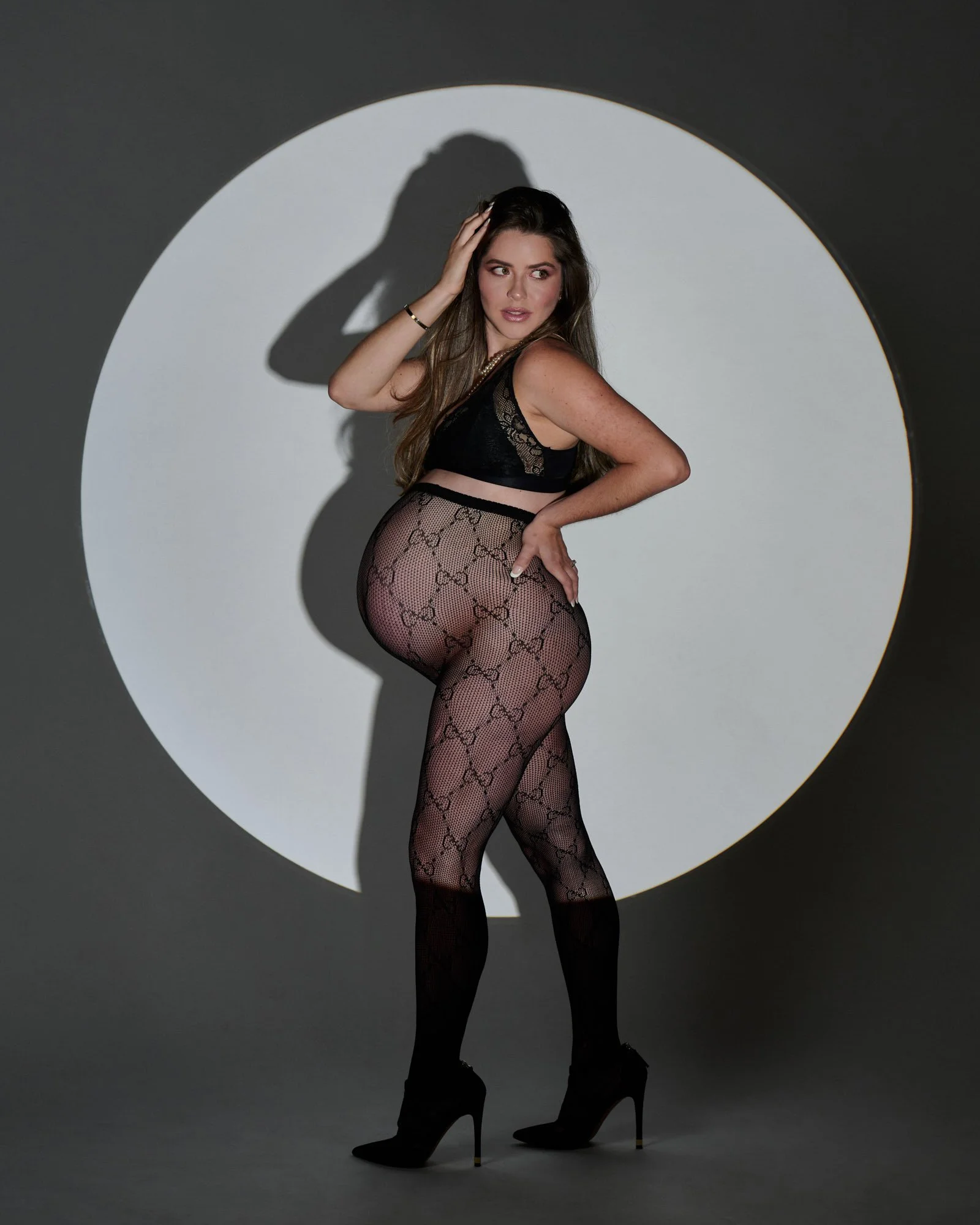 Woman wearing black lace top and patterned tights posing in front of a circular white spotlight projection.