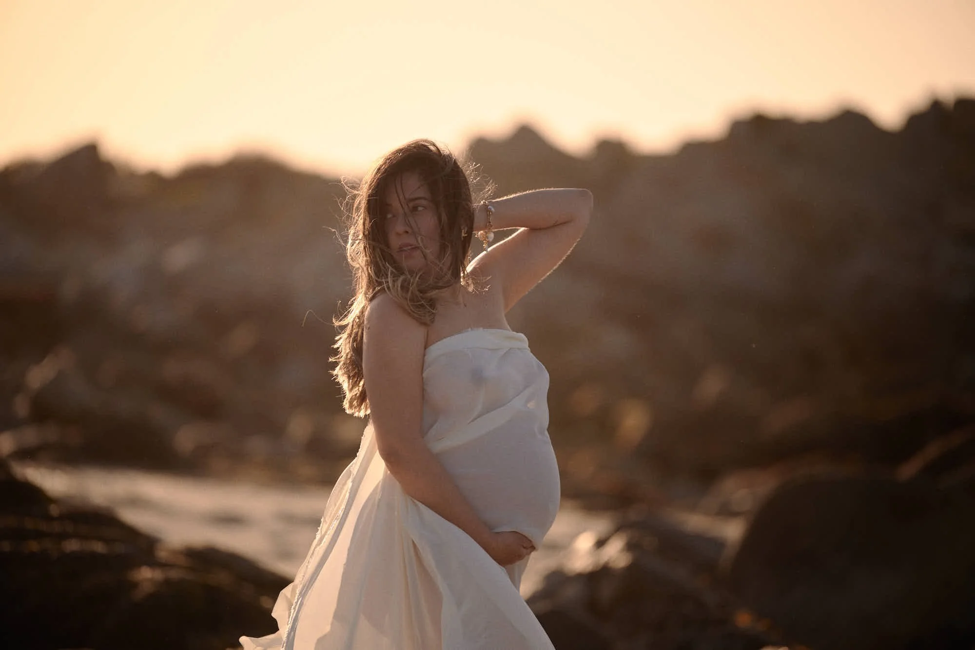 Strapless white gown at golden hour on the rocks, hand in her hair and surf glowing behind.