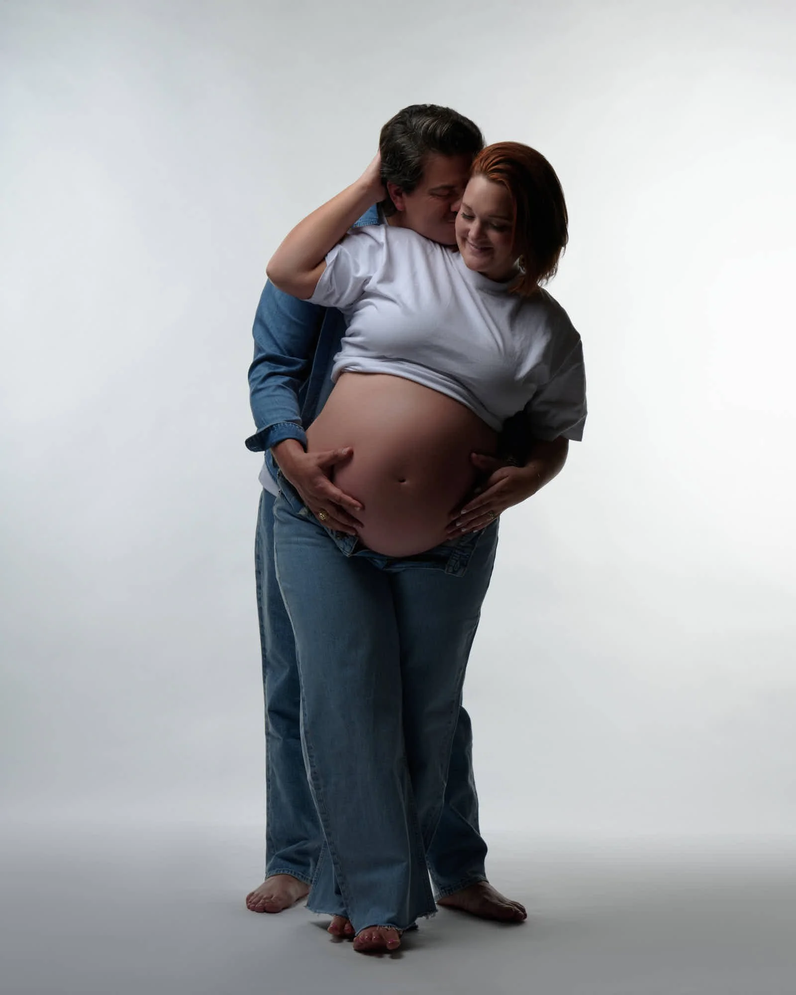 Partner kisses the mother-to-be's cheek from behind, hands stacked on her bare bump.