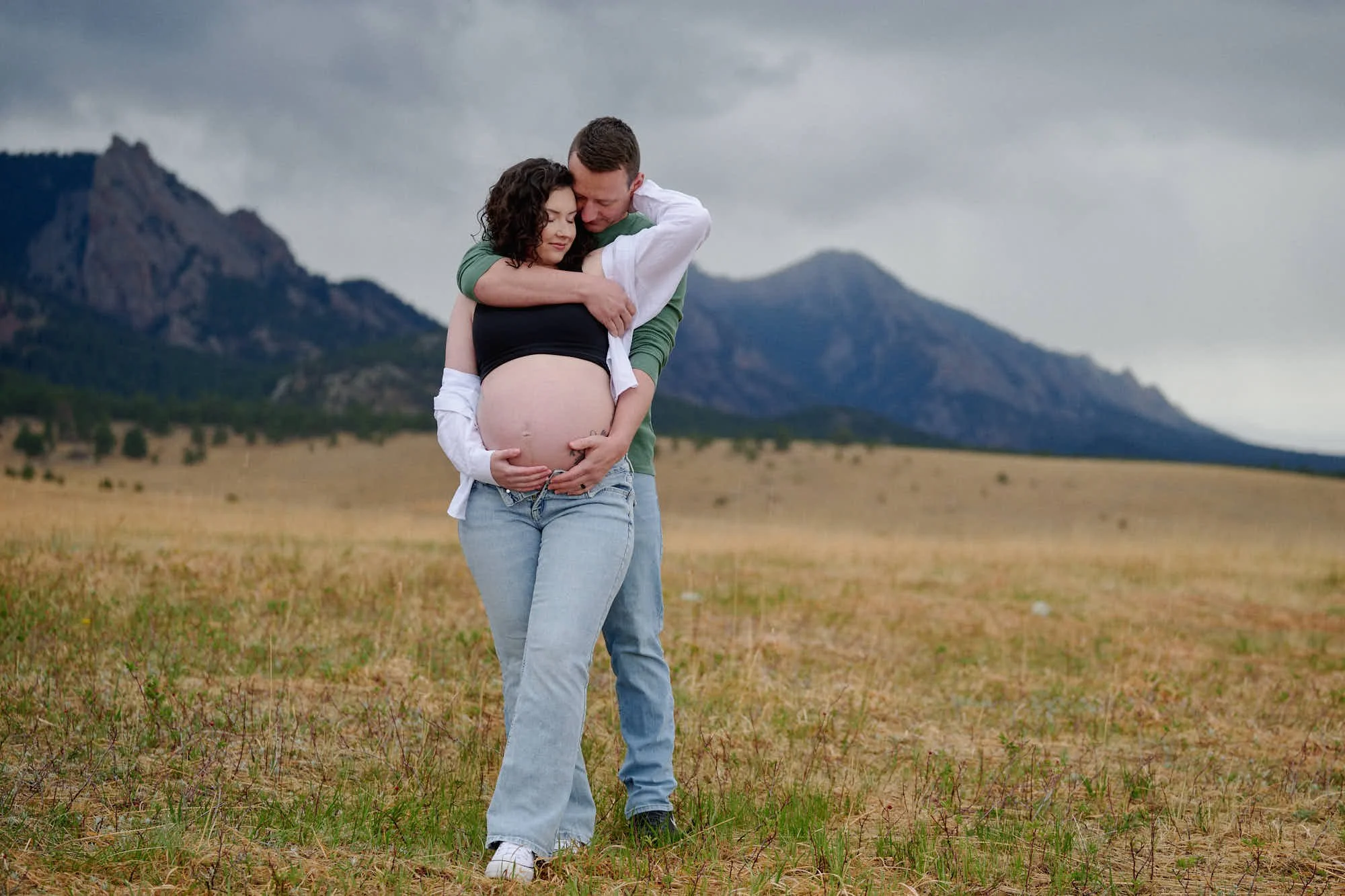 He hugs her from behind under a brooding sky, open white shirt framing the bump.