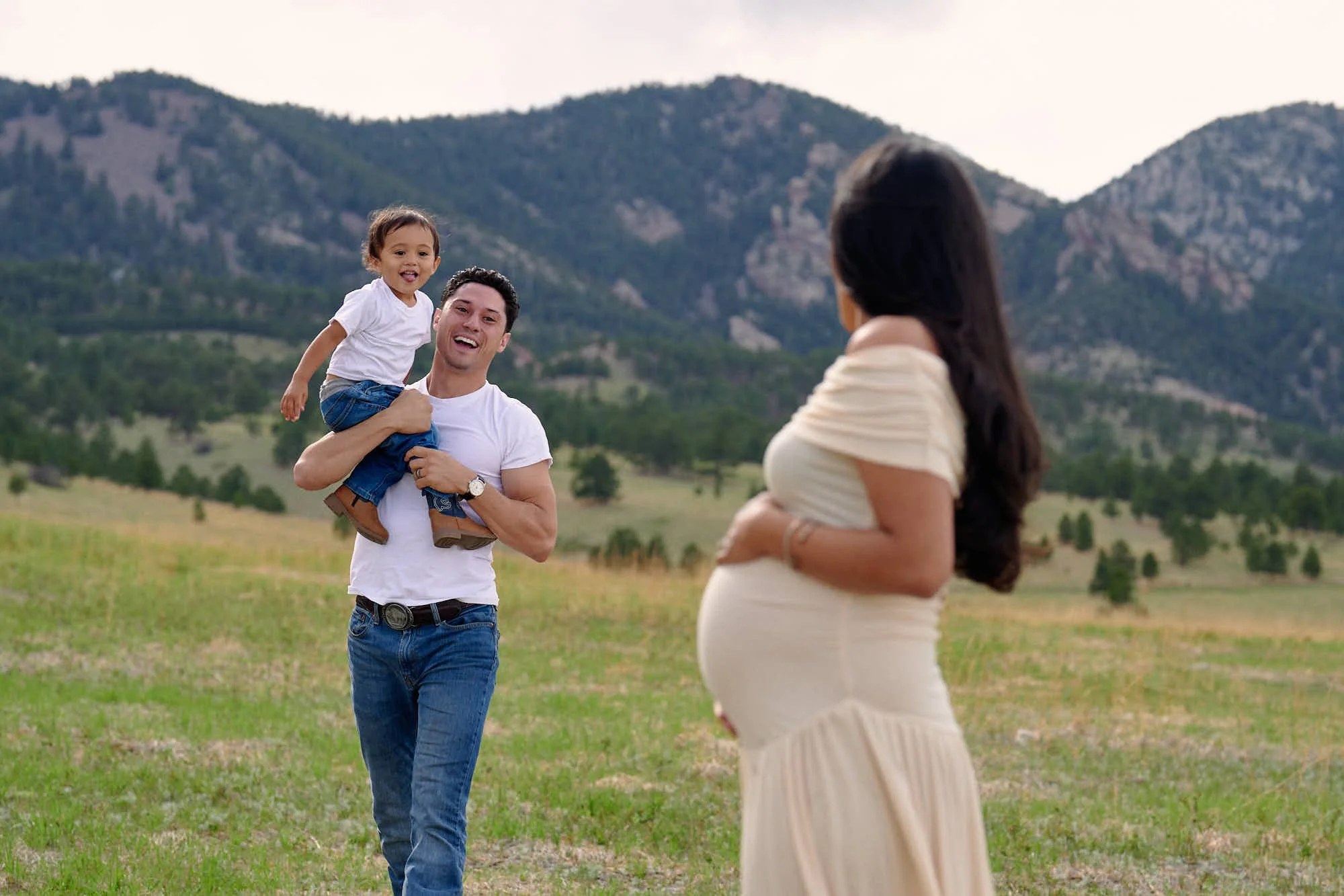 Dad lifts their toddler high while mom laughs in a cream ruffled gown, mountains all around.